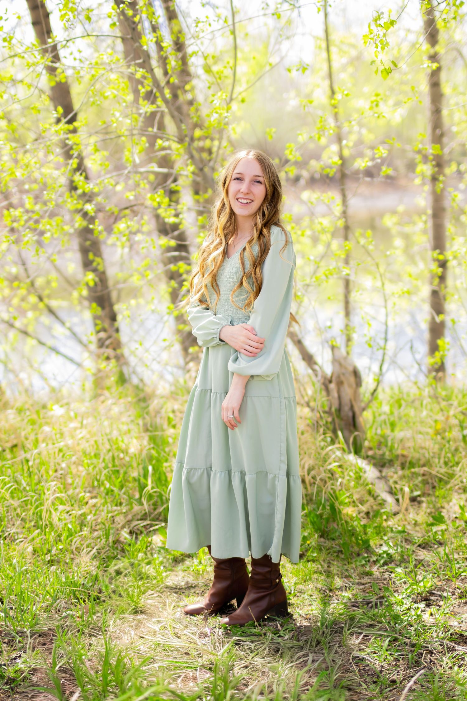 Young woman in mint green maxi dress and brown boots smiling outdoors among spring trees near water.
