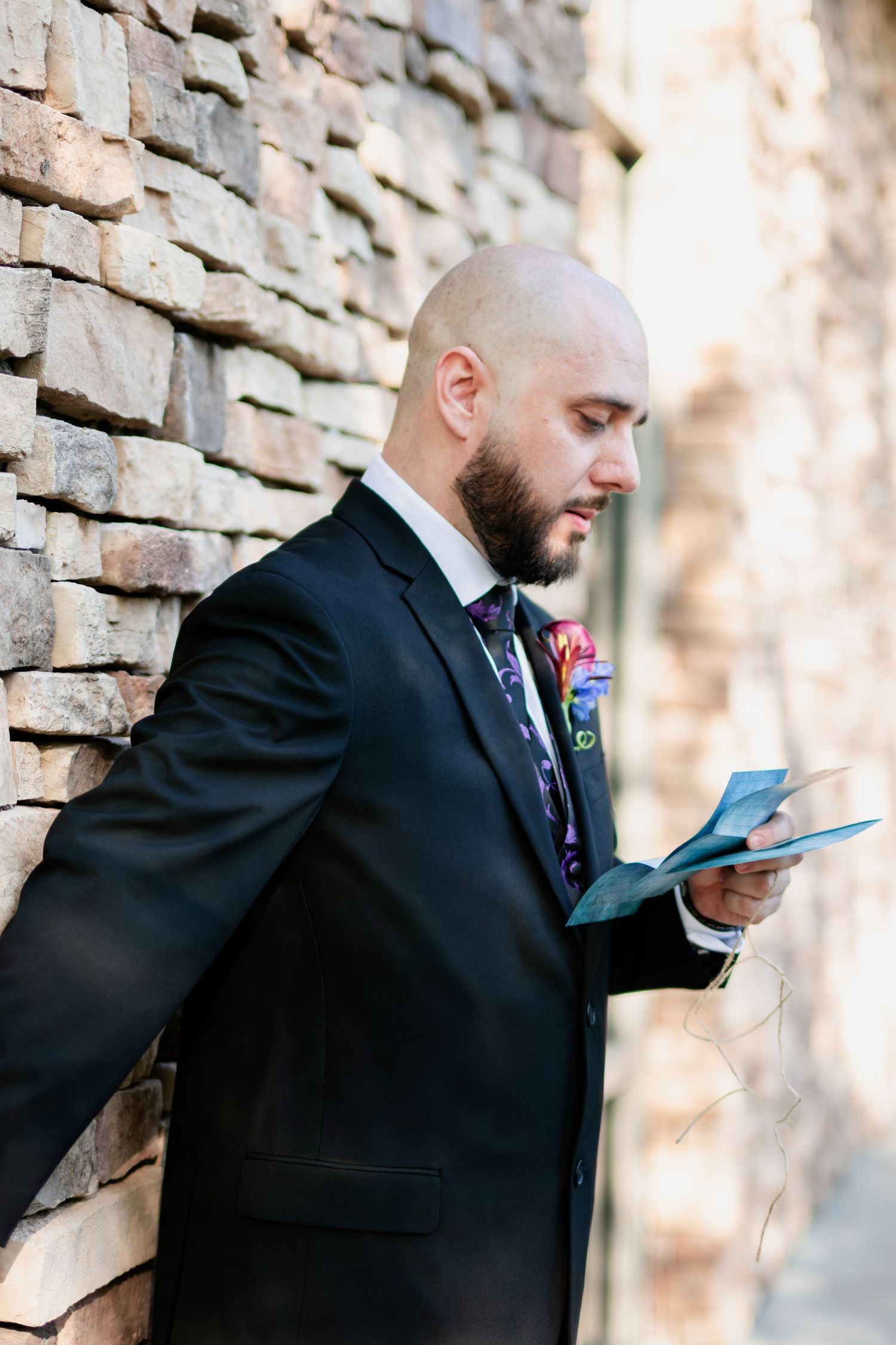 Bald groom in black suit reading vows card leaning against stone wall with colorful boutonniere.