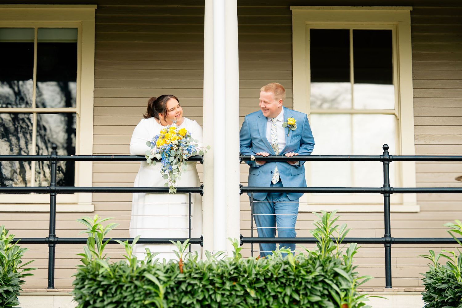 A couple stands together on a porch with black railings and green plants in the background.