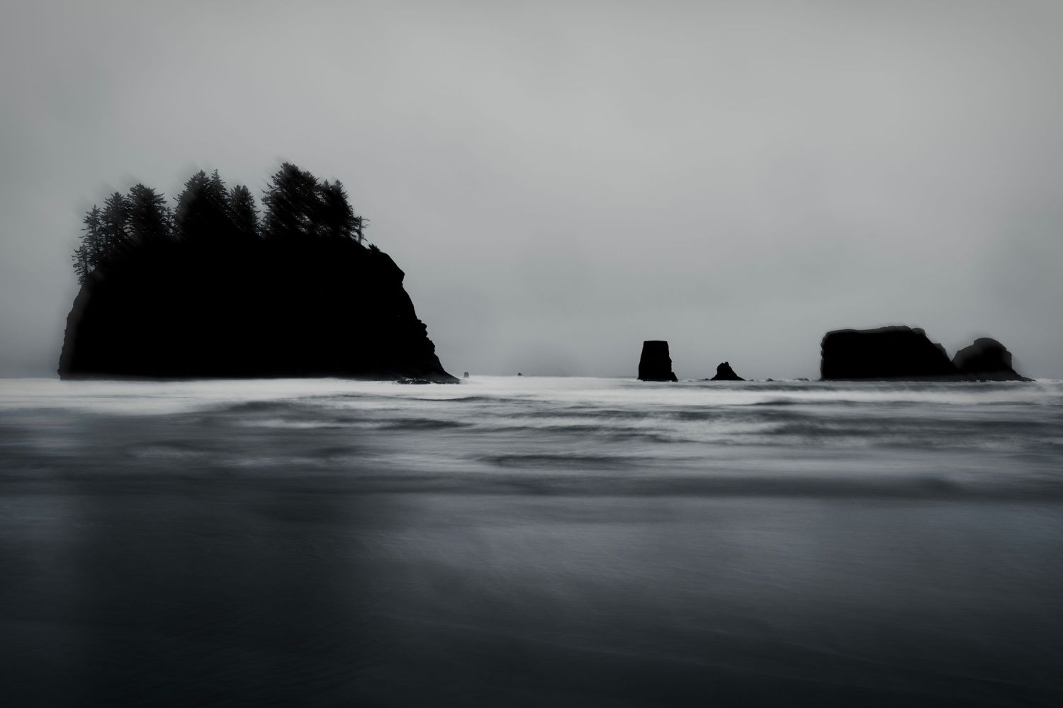 Dramatic black and white coastal seascape showing rugged rock formations and crashing waves along a misty shoreline.