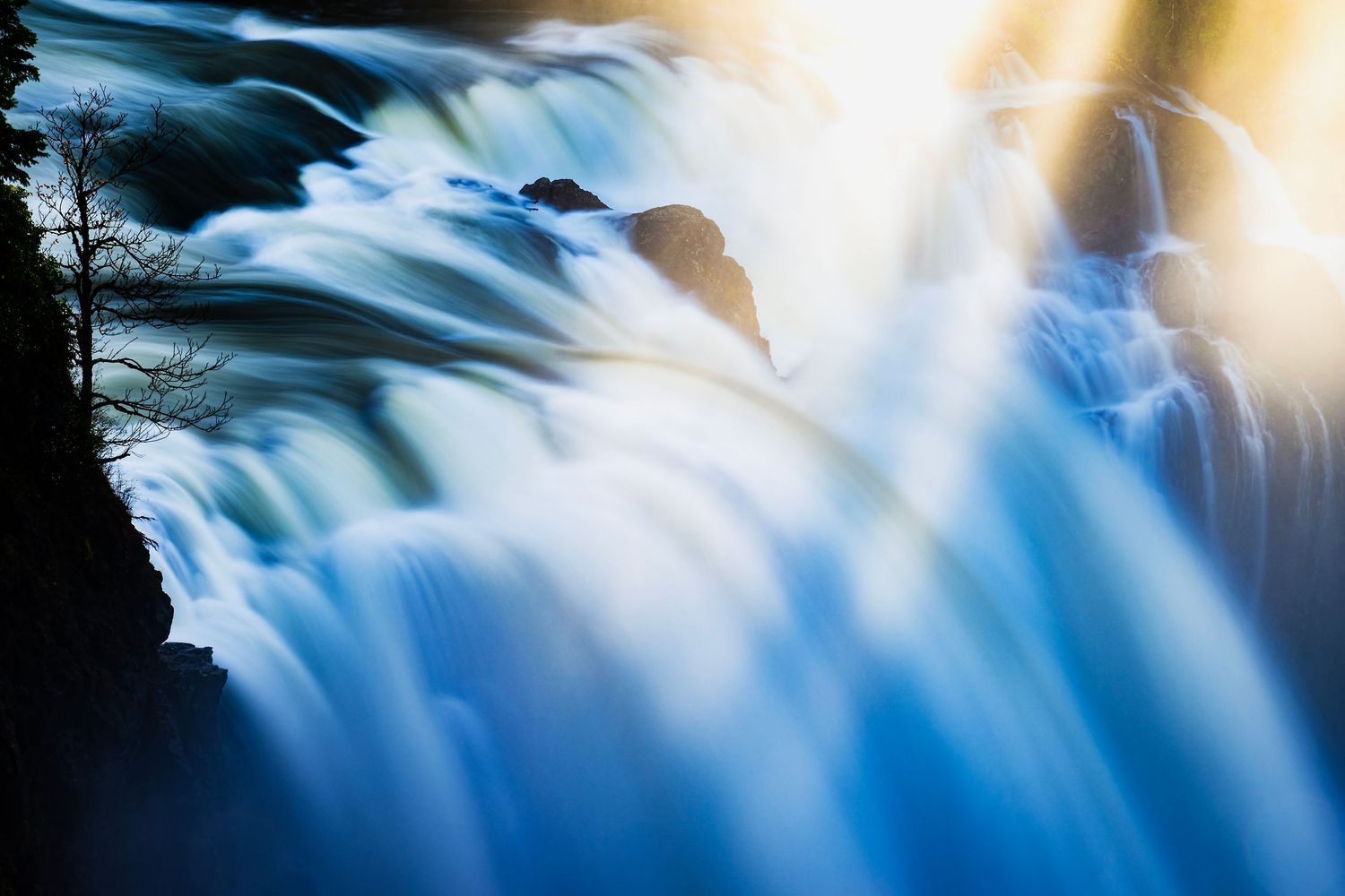Scenic waterfall cascading over rocks with sunlight streaming through mist creating a dramatic natural landscape.