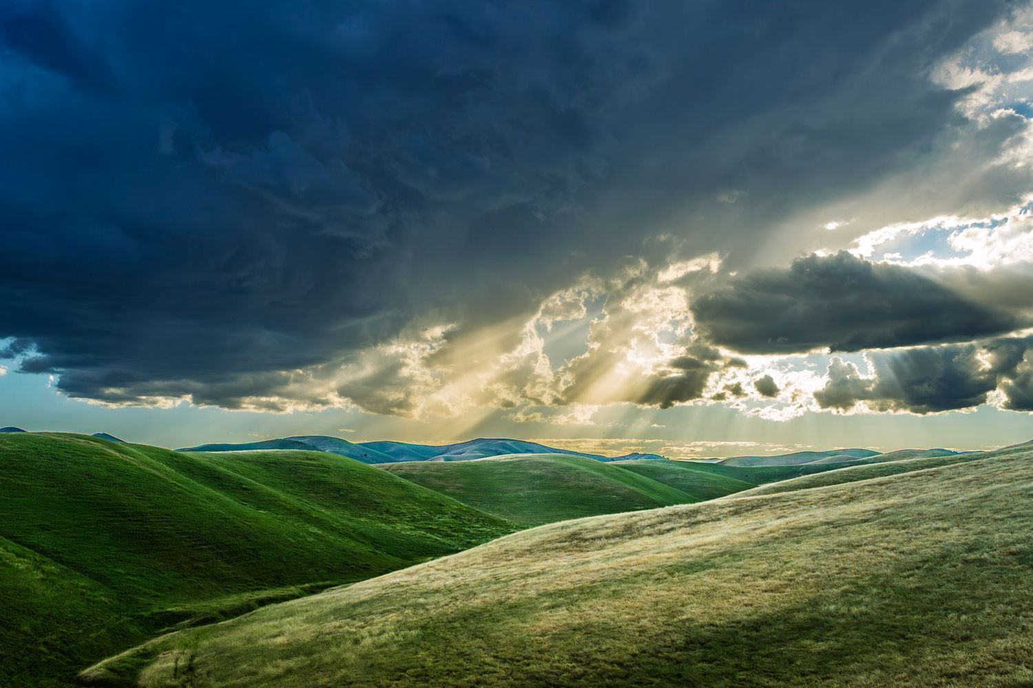 Rolling green hills under dramatic storm clouds with sunbeams breaking through, illuminating the lush landscape.