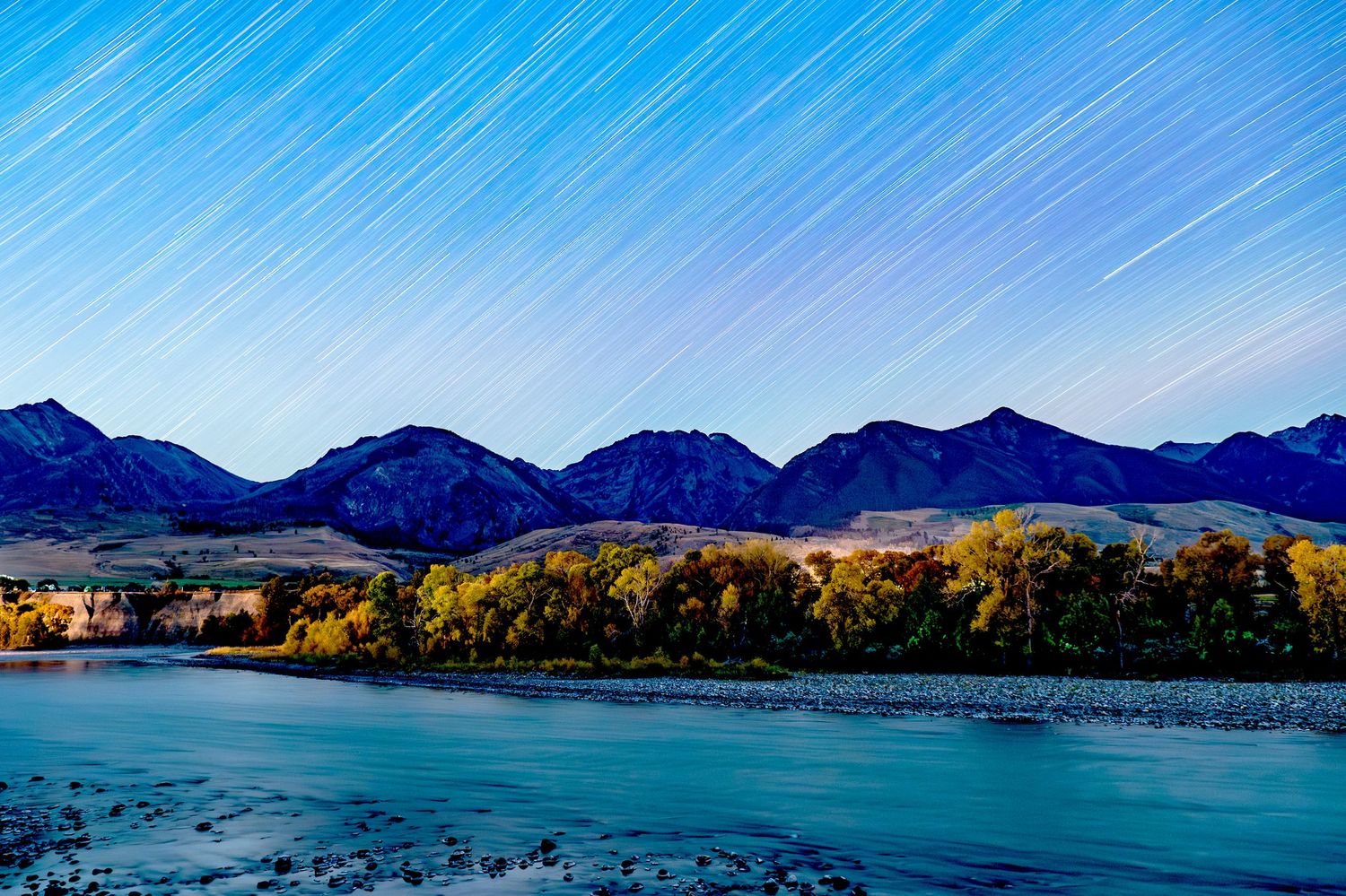 A panoramic mountain landscape view with snow-capped peaks reflected in a tranquil lake under blue skies.