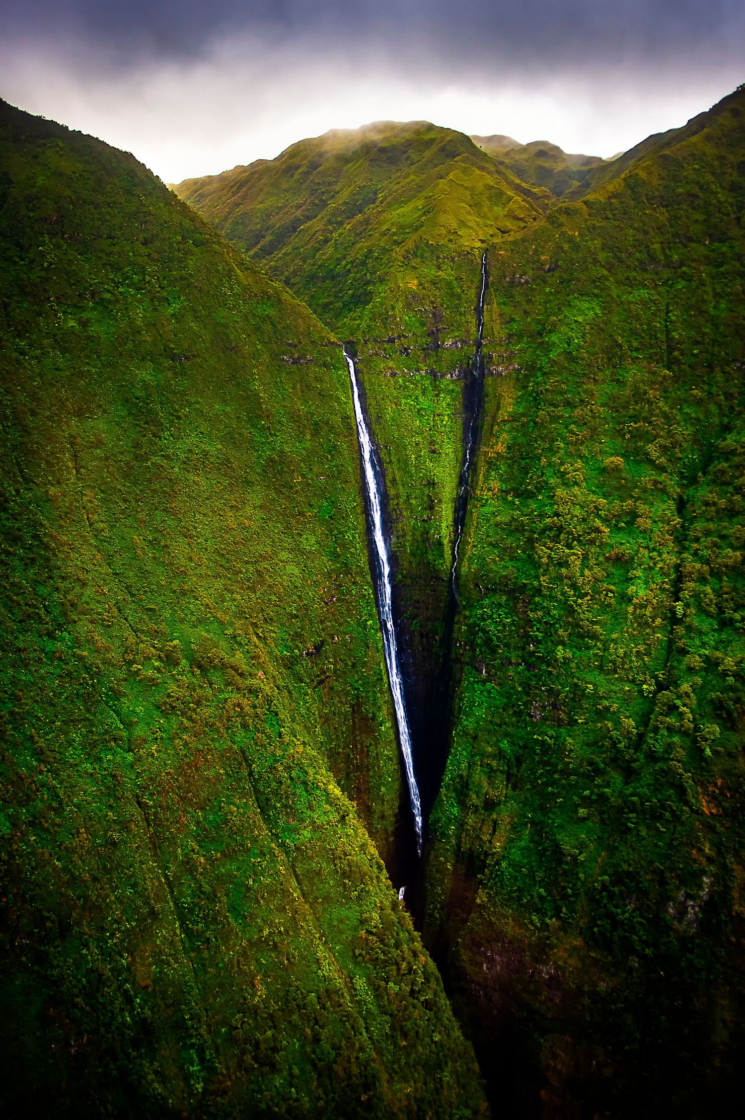 Tall cascading waterfall plunges down steep green mountainside in Hawaii's lush tropical valley under moody skies.