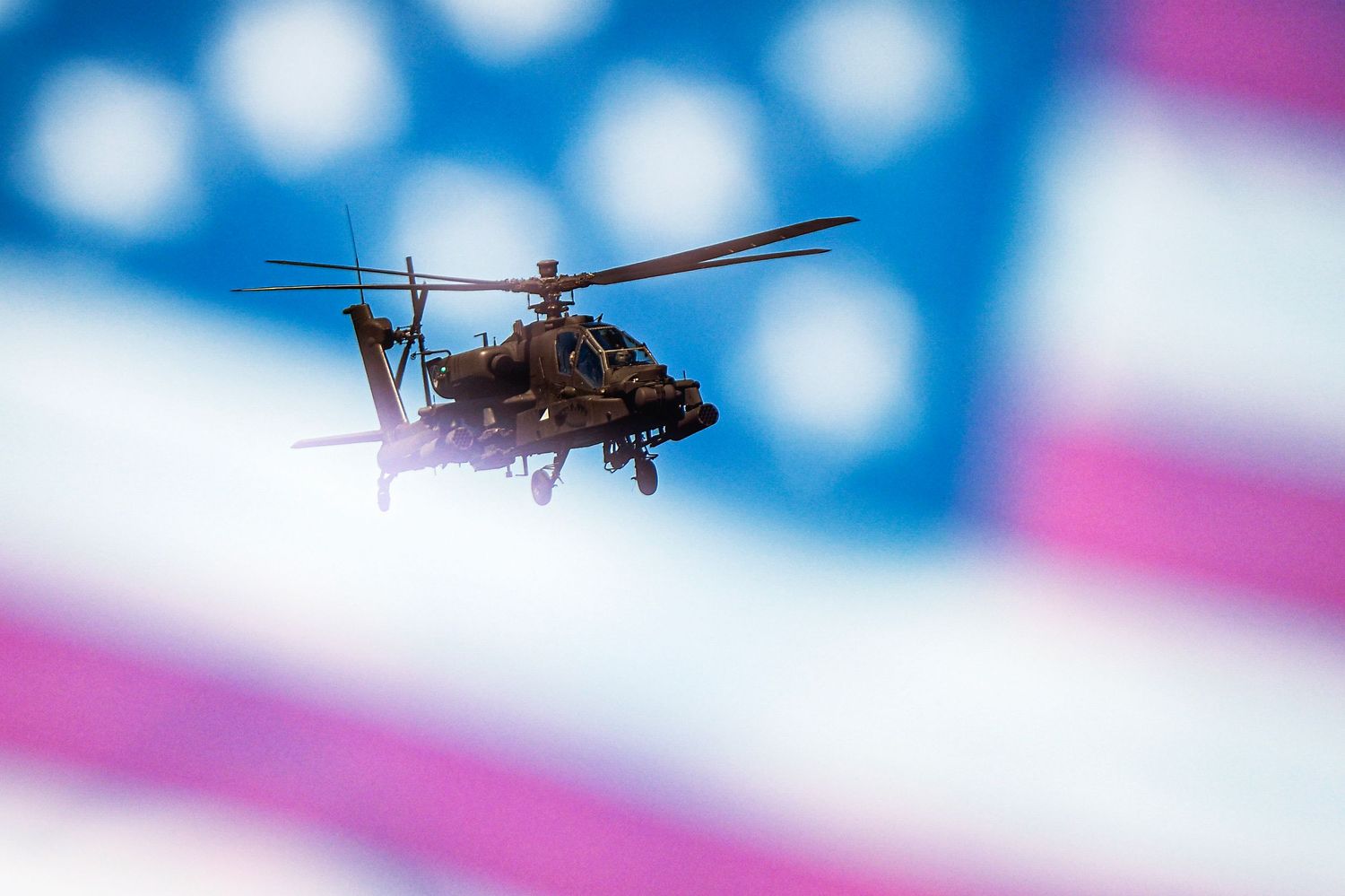 Military Apache helicopter flying in front of a blurred American flag backdrop.