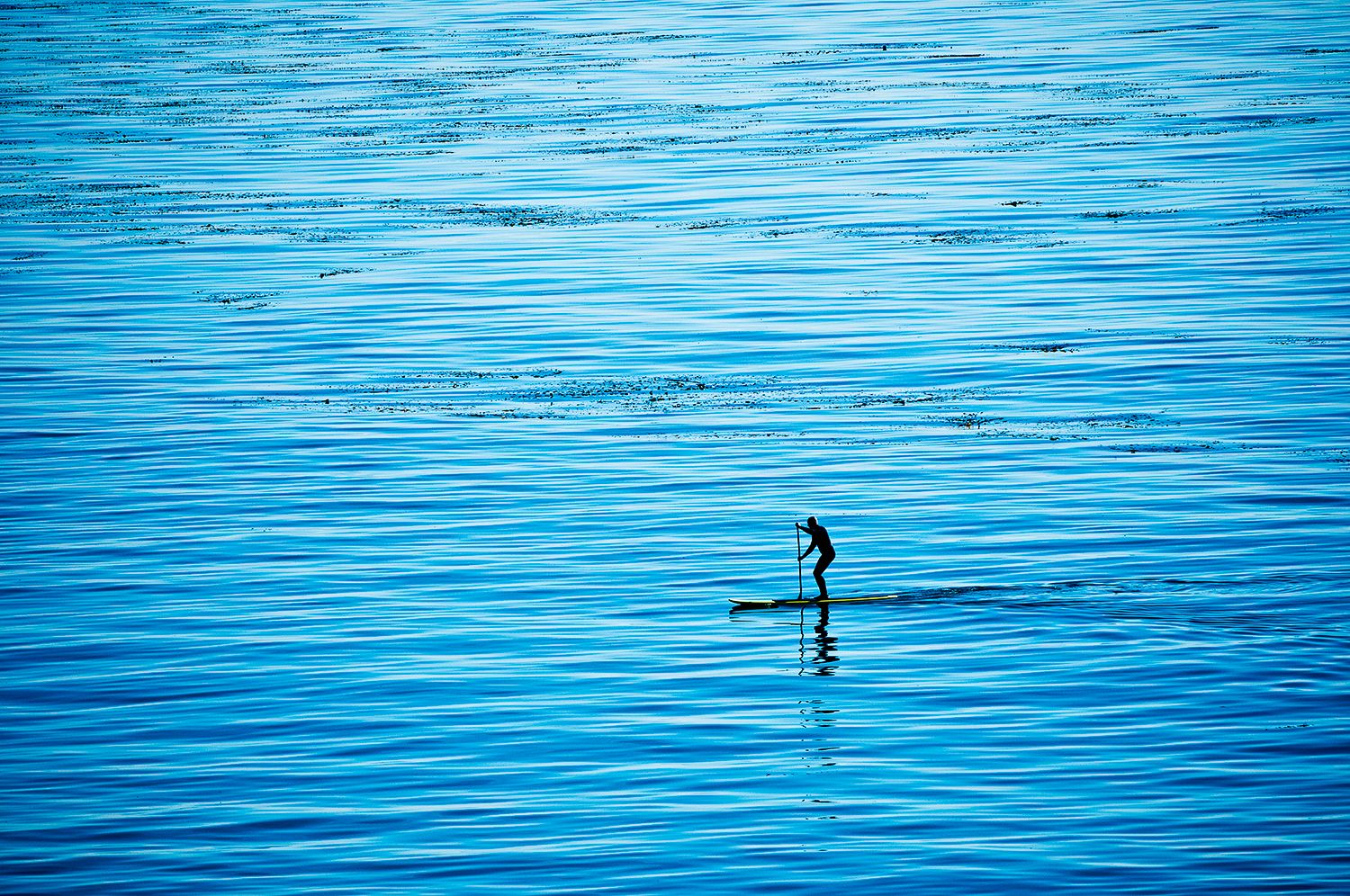 A lone paddleboarder stands on calm blue waters creating a minimalist silhouette against the rippling ocean surface.