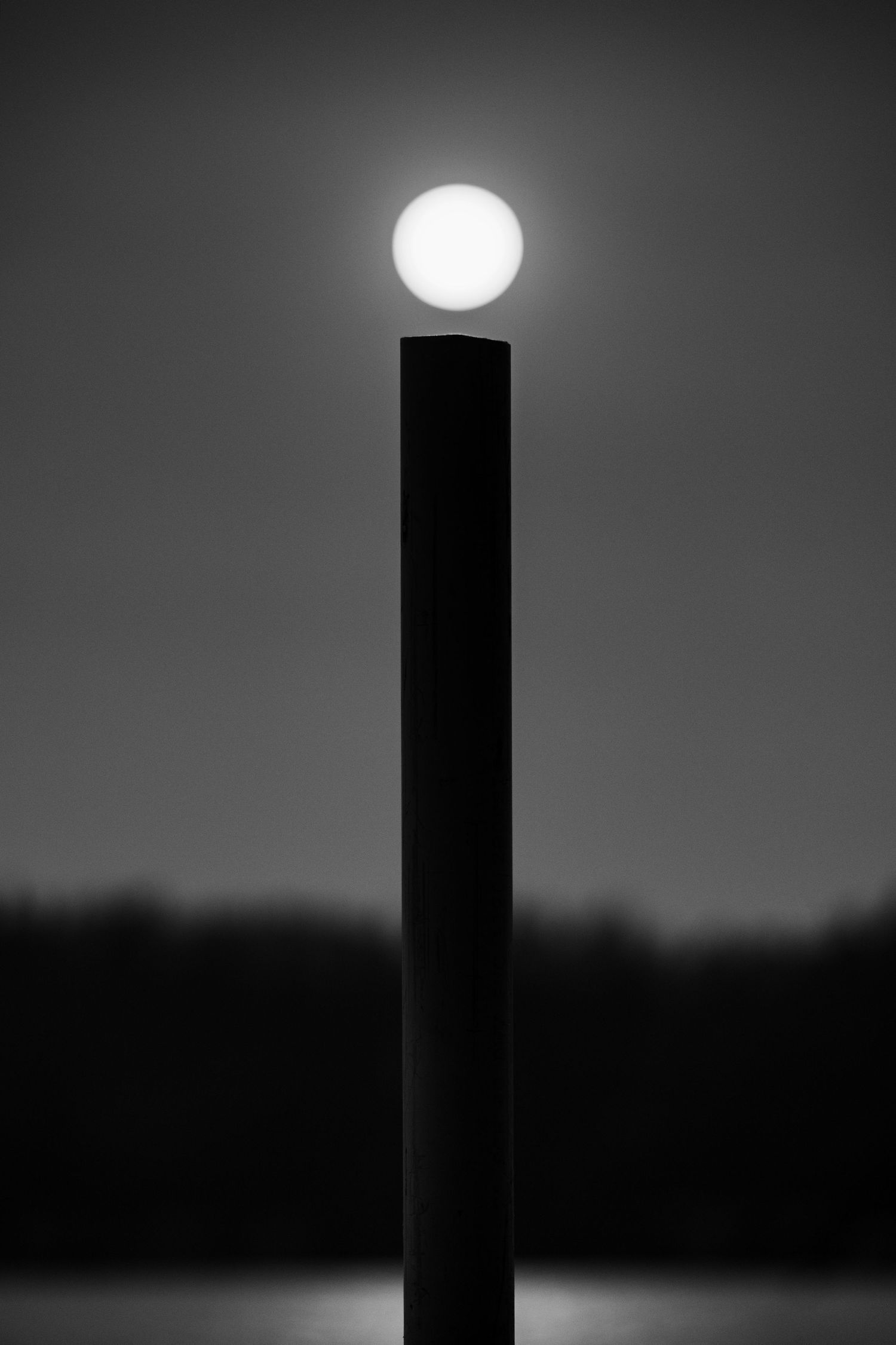 Black and white photo of a glowing spherical light atop a dark pole against a moody night sky.