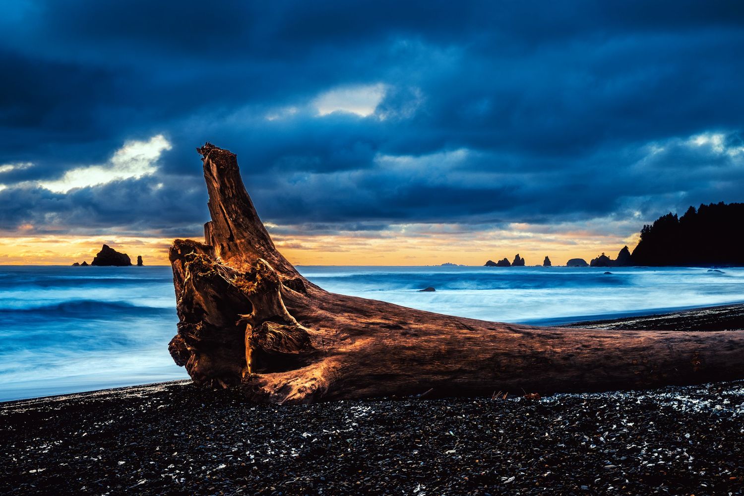 A large driftwood log rests on a black sand beach at sunset with dramatic blue storm clouds overhead.