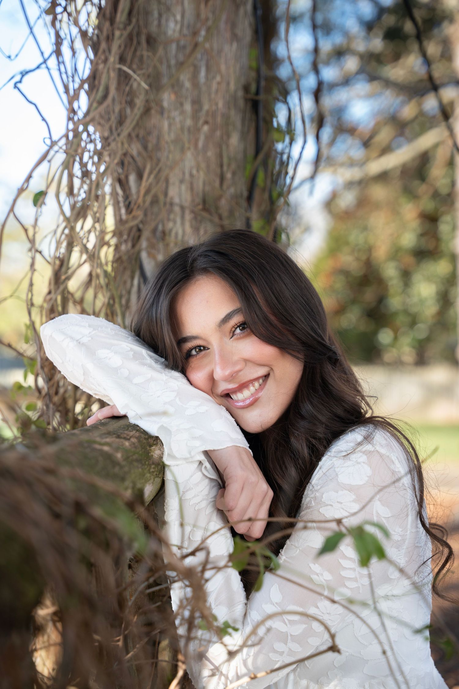 A person in a white puffy winter jacket poses playfully near tree branches on a sunny autumn day.