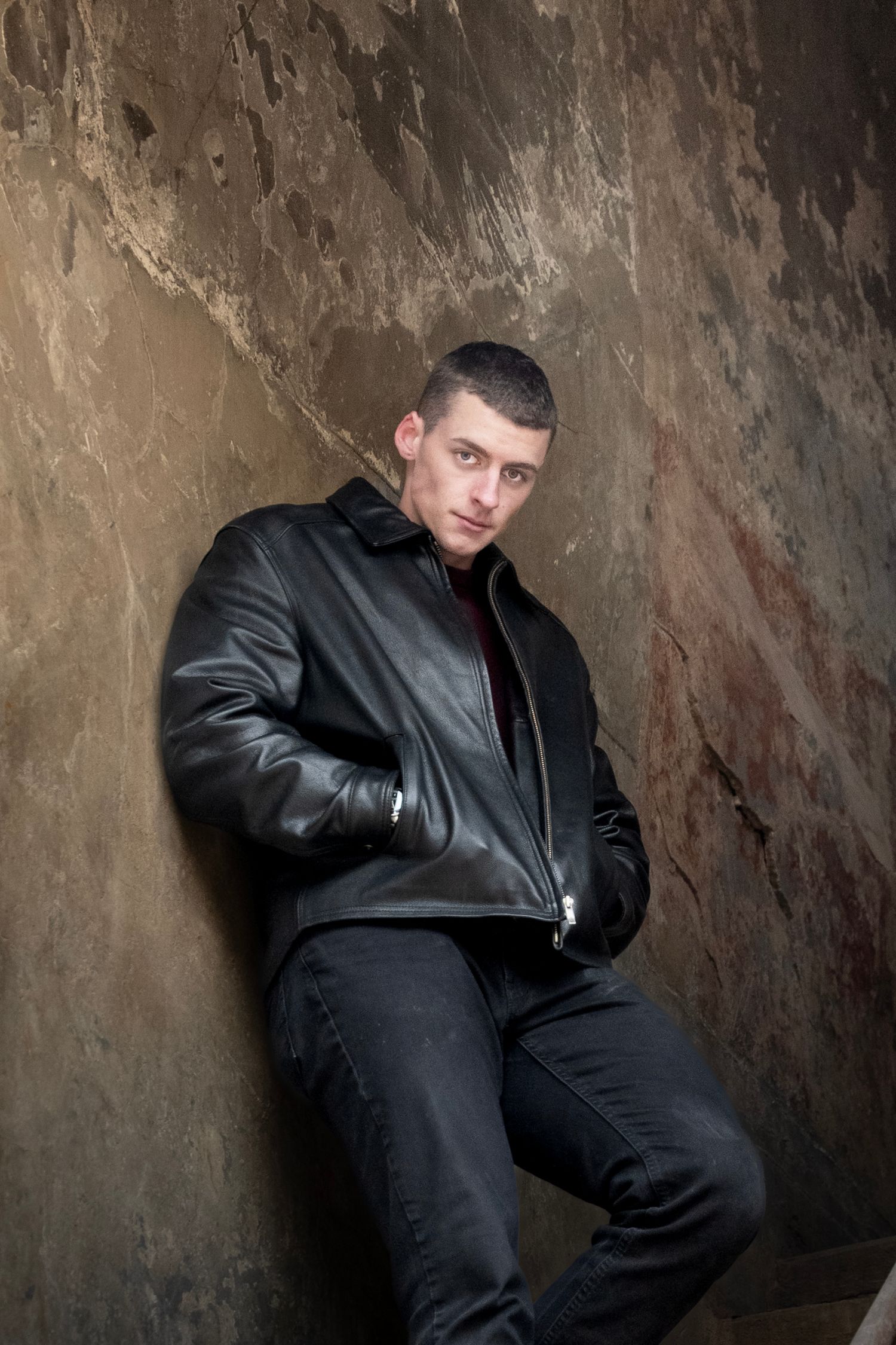 Person wearing black leather jacket poses against textured stone wall in dramatic lighting.