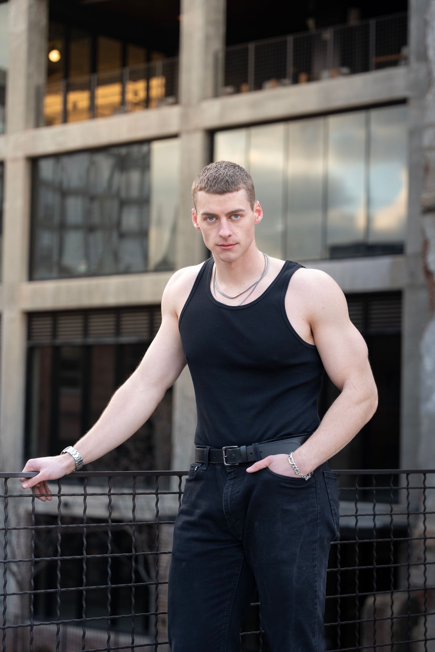 Athletic person in black tank top and dark jeans poses casually against railing of urban building.