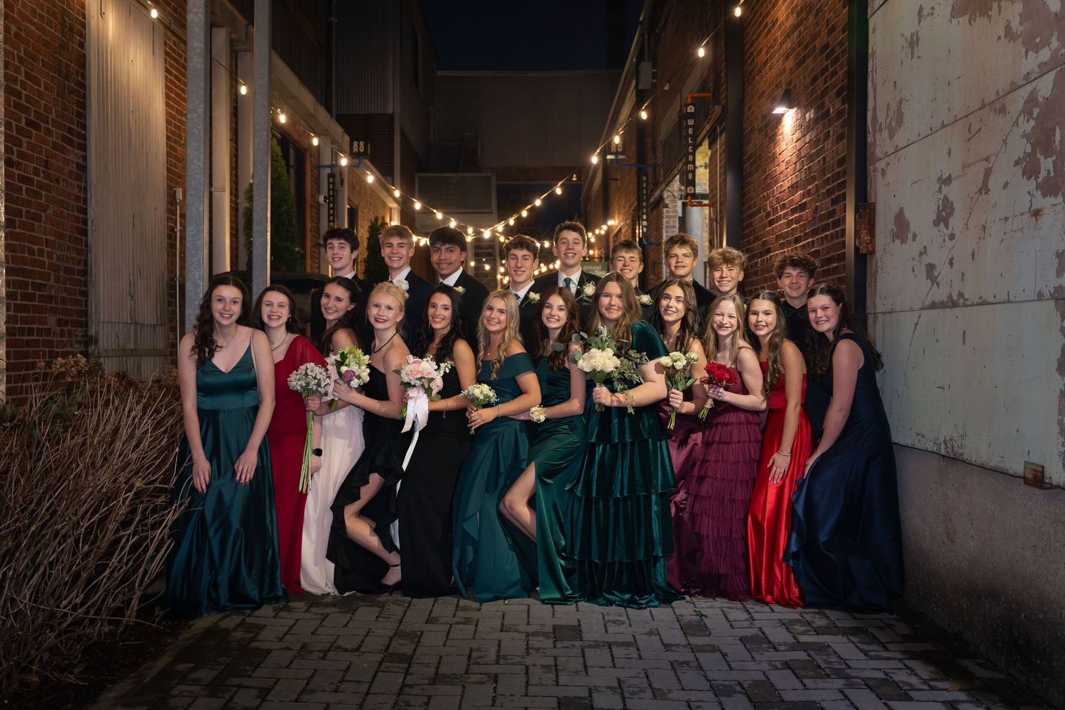 A group in formal evening gowns poses together under string lights in an urban alleyway at night.