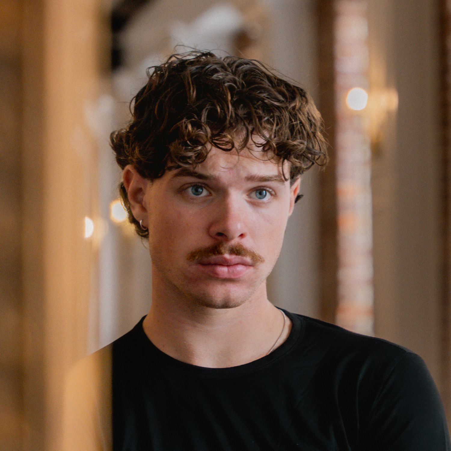 Portrait of a young man with curly brown hair and a mustache wearing a black shirt, standing in a warmly lit hallway.