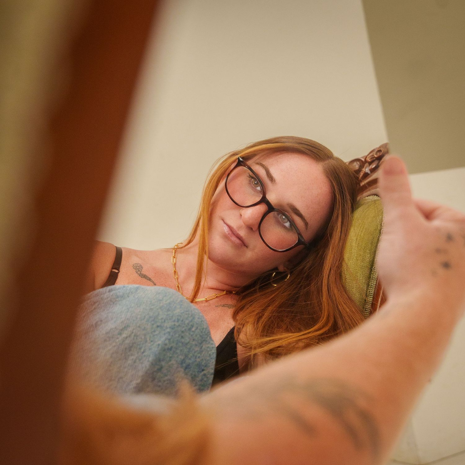 Young redhead woman with glasses and tattoos relaxing on a couch, viewed from a low angle perspective.