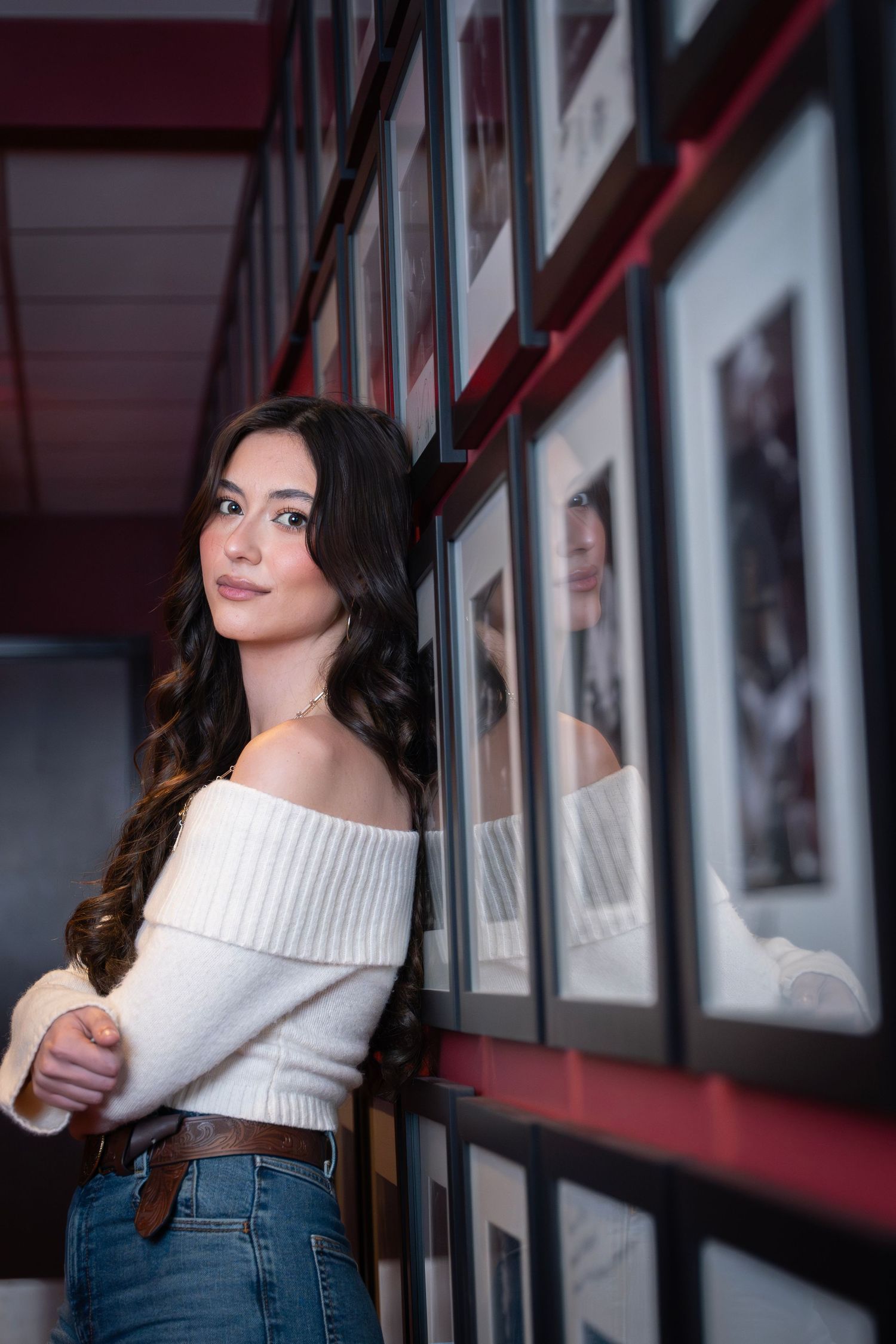 A person in a white off-shoulder sweater and jeans poses in a hallway with framed artwork on the wall.