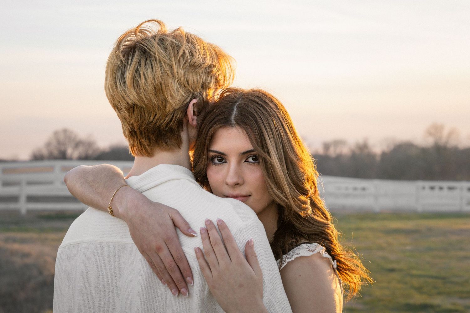 A couple embraces in a golden sunset light on a rural field with fencing in the background.