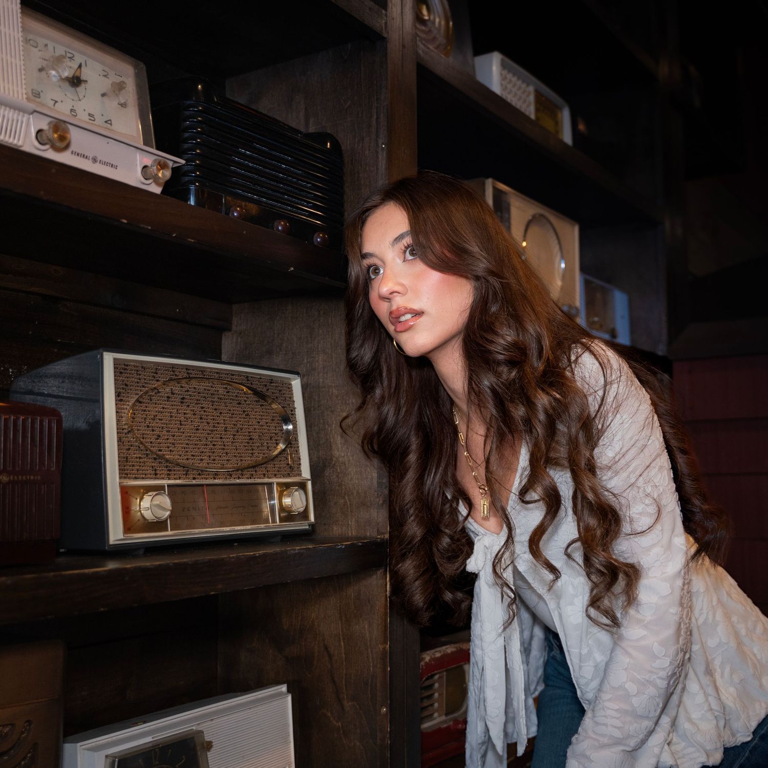 A person in a light colored jacket poses in a dimly lit interior space with wooden beams and shelving.