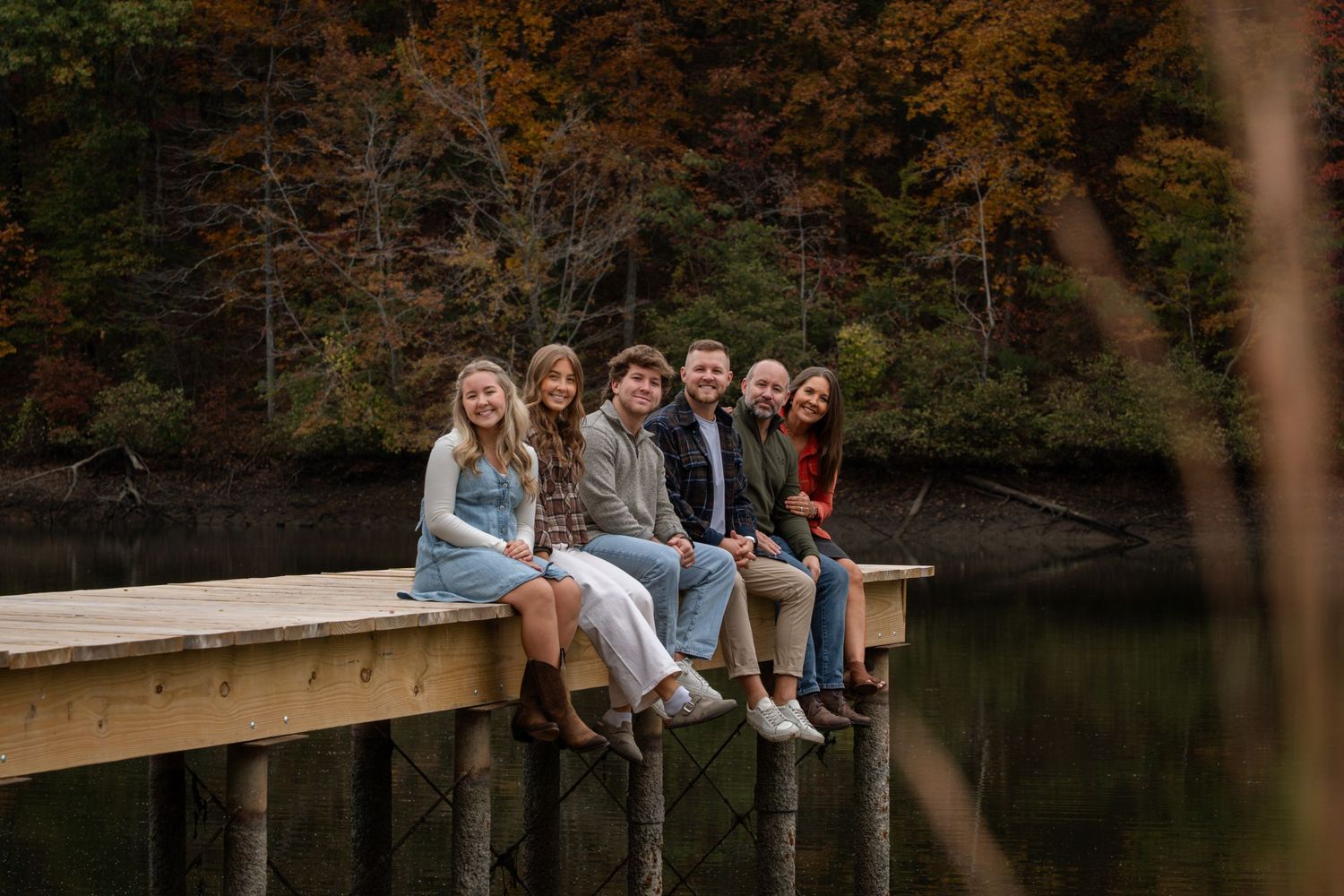 People sit together on a wooden dock overlooking a serene lake surrounded by autumn trees.