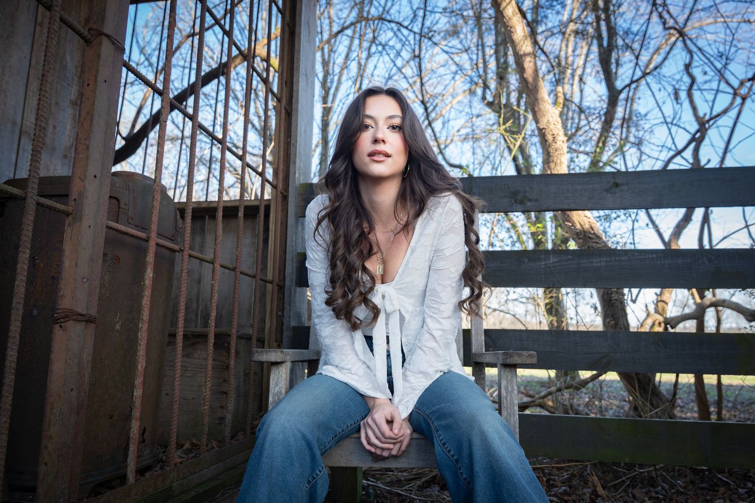 Person with long dark hair sits on wooden deck in winter setting wearing casual attire and looking contemplative.
