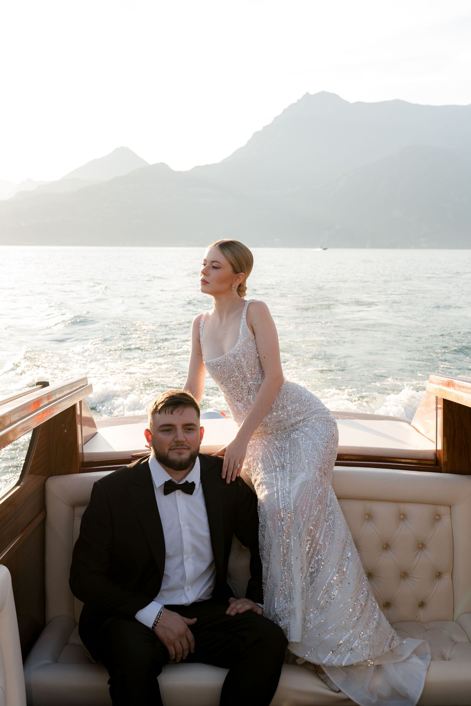 Bride in sequin gown and groom in tuxedo pose on a luxury yacht with mountain backdrop at sunset.