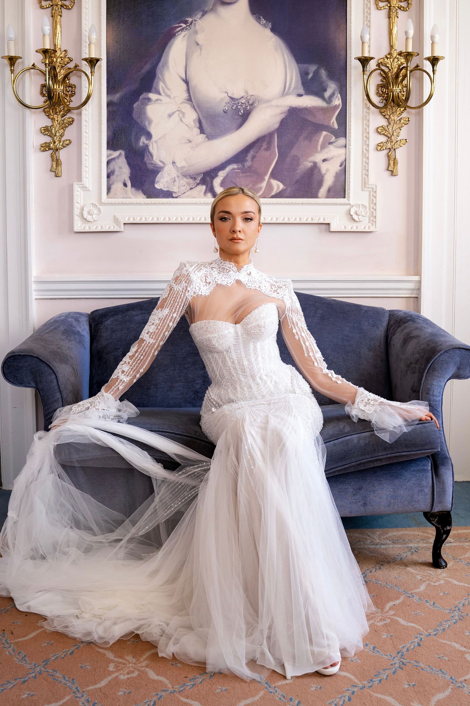 Elegant bride in structured lace wedding gown seated on blue velvet sofa in ornate classic room.