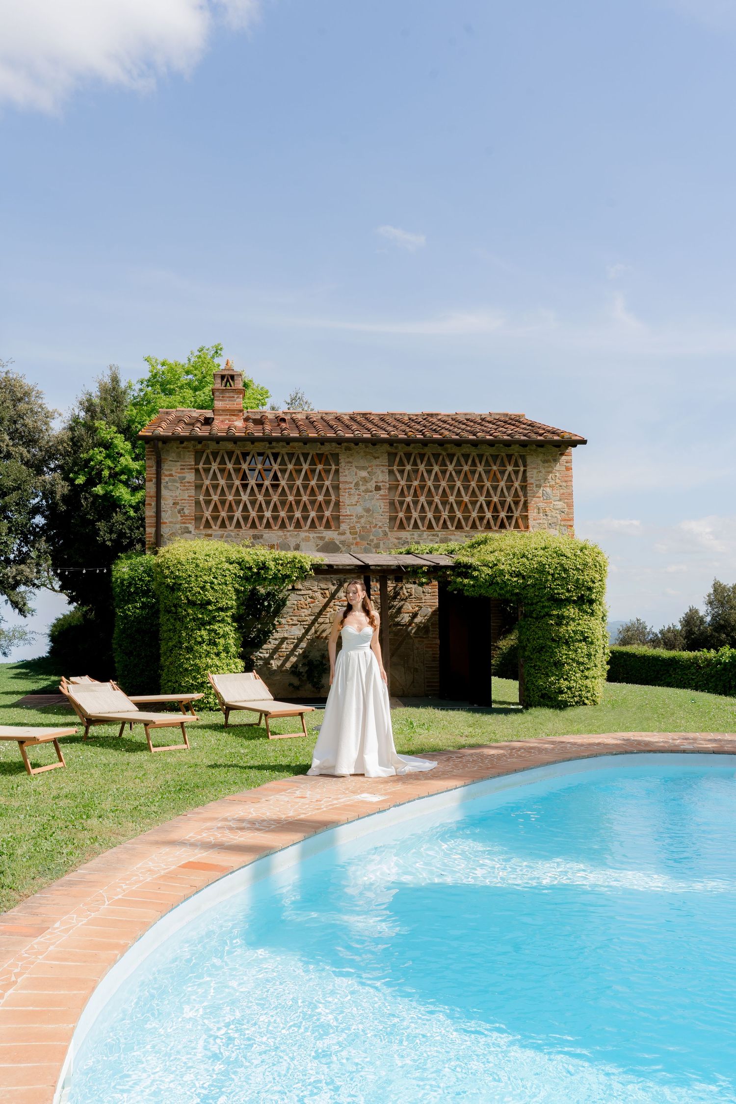 Elegant bride in white gown stands beside a sparkling pool at a rustic Italian villa wedding.