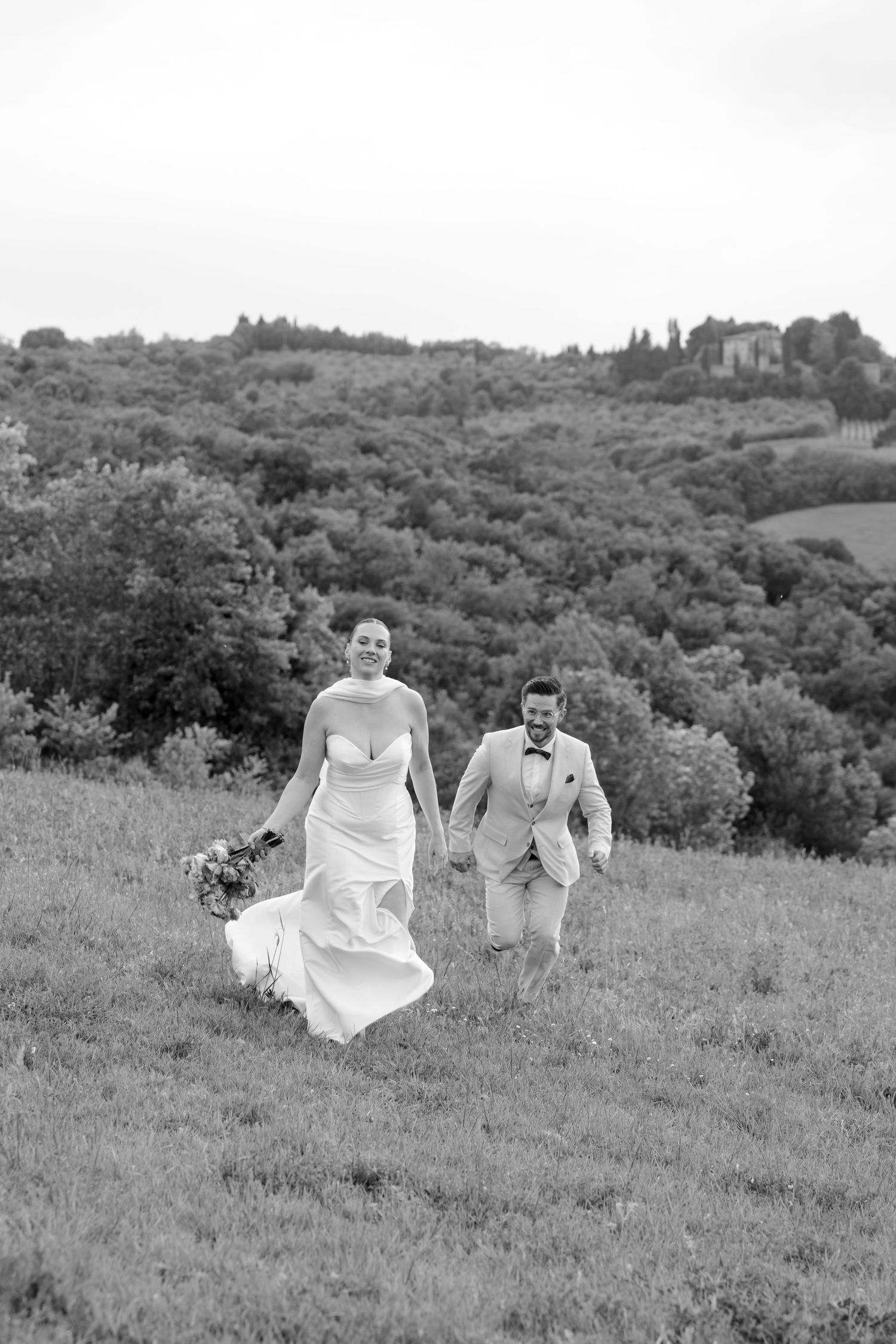 Black and white photo of laughing bride and groom running through a Tuscan hillside meadow.