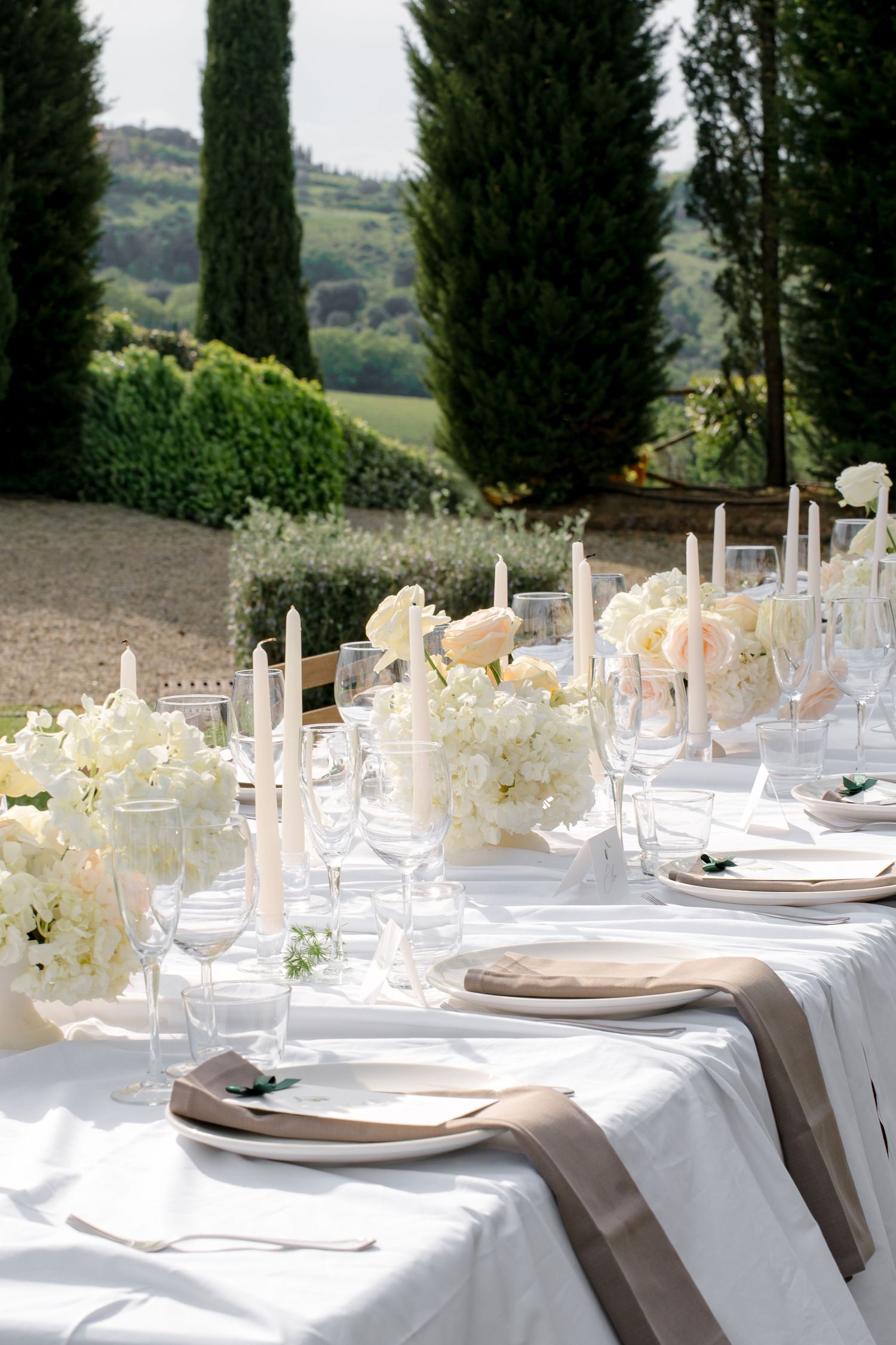 Elegant outdoor wedding reception table with white florals, tapered candles, and Tuscan cypress backdrop.