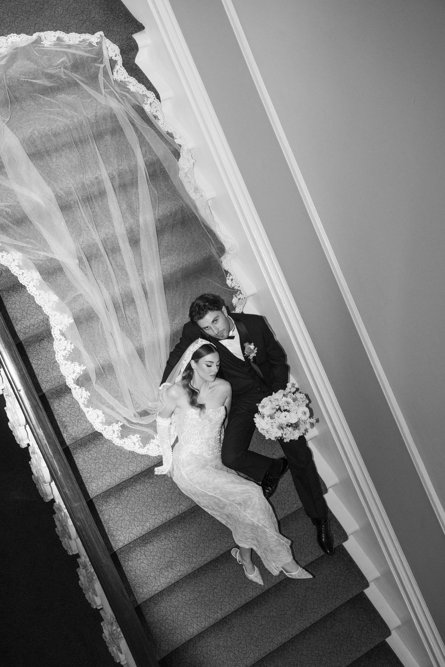Aerial black and white wedding photo of bride and groom on staircase with flowing lace veil.