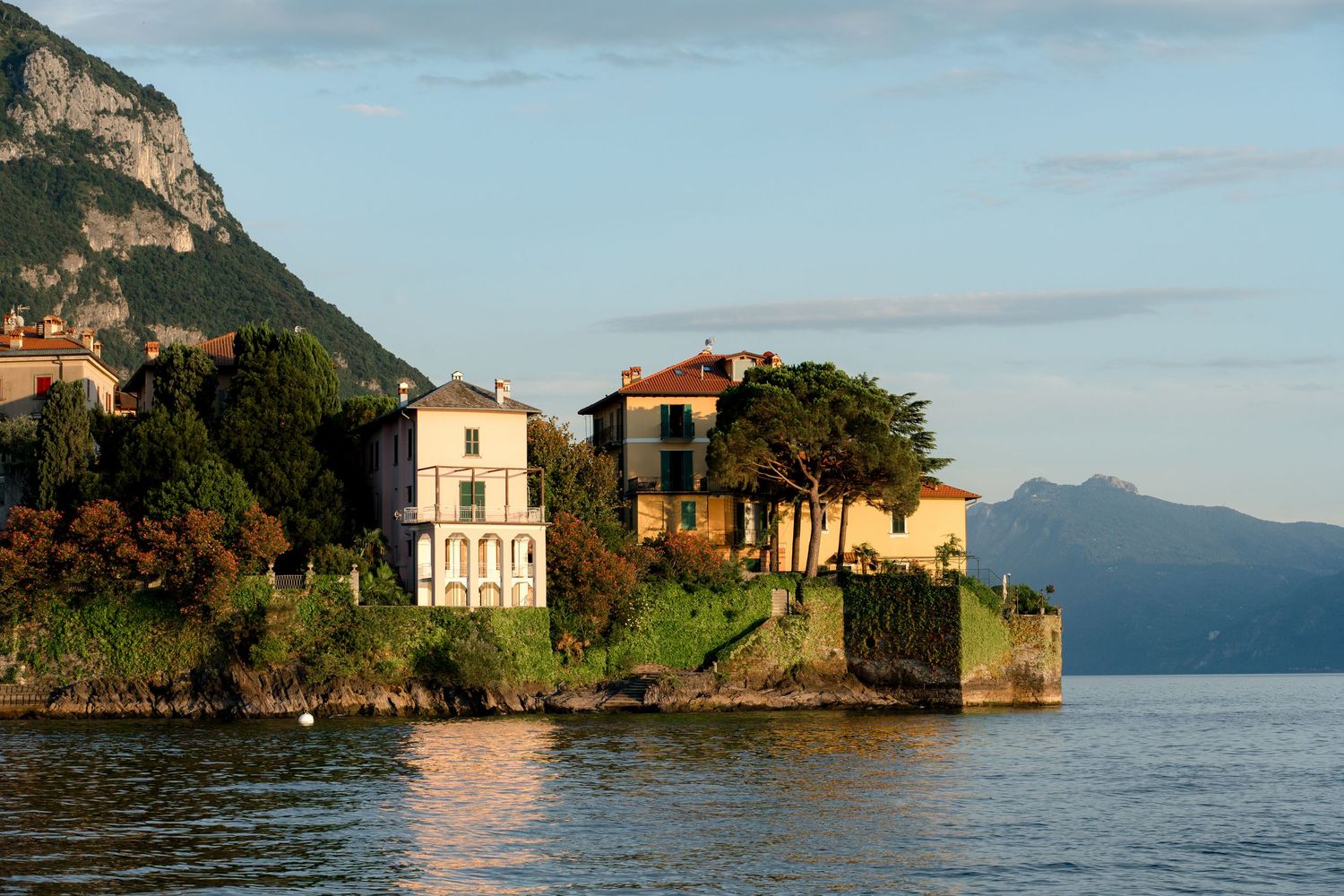 Elegant lakeside villas nestled among lush trees on Lake Como, Italy, with mountain backdrop at golden hour.