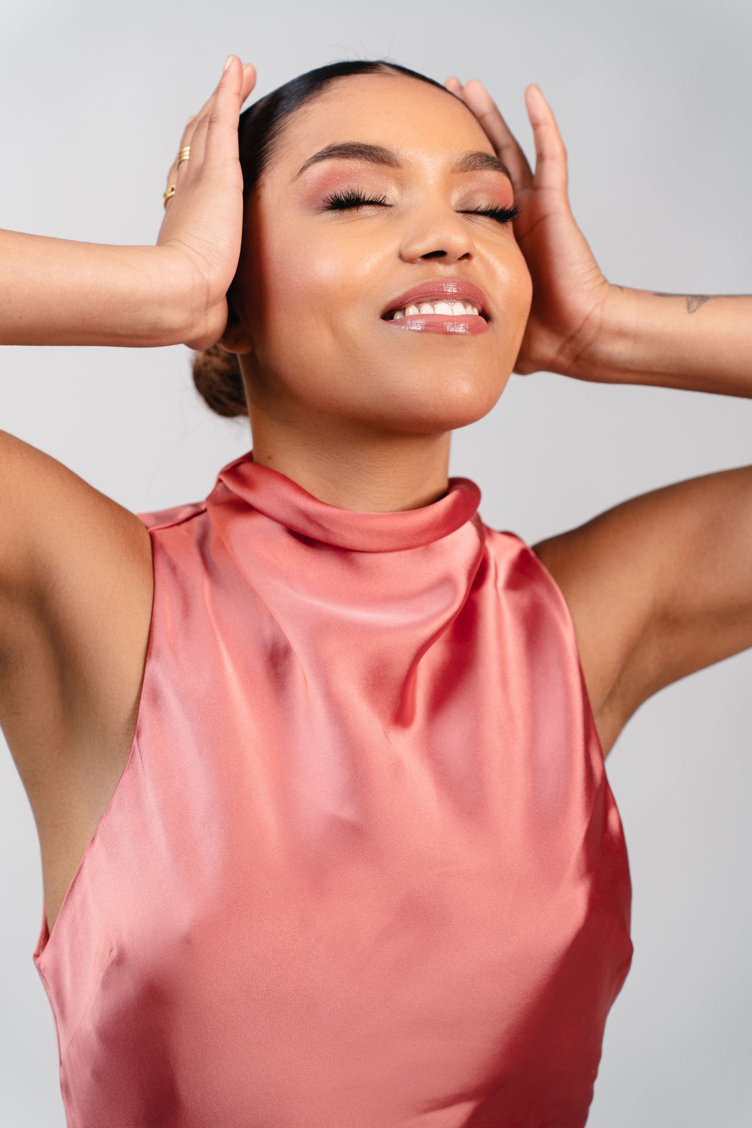 Woman in a pink satin sleeveless dress smiling with eyes closed, hands framing her face against a gray background.