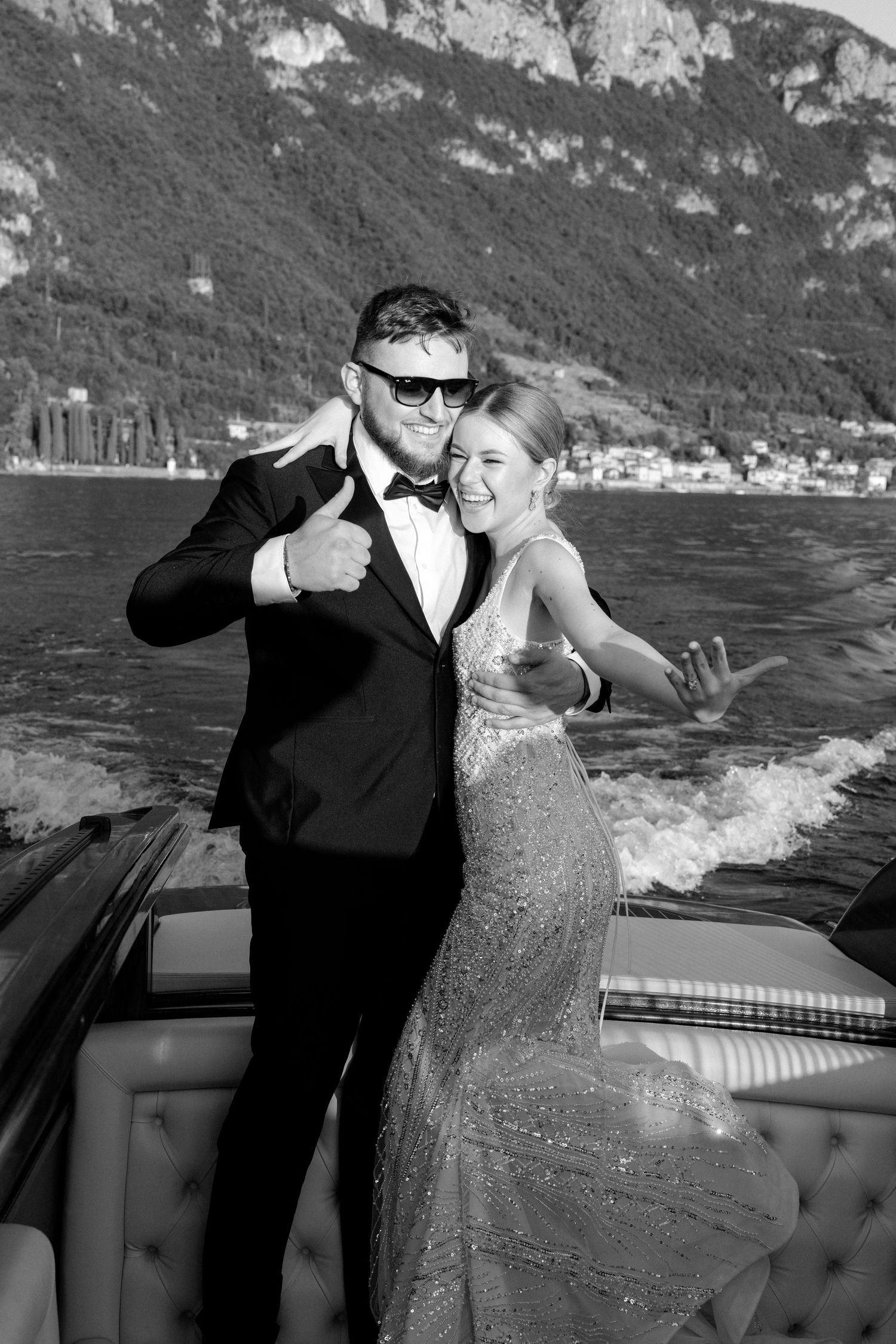 Elegant couple in formal attire posing on a luxury boat with mountain lake backdrop in black and white.