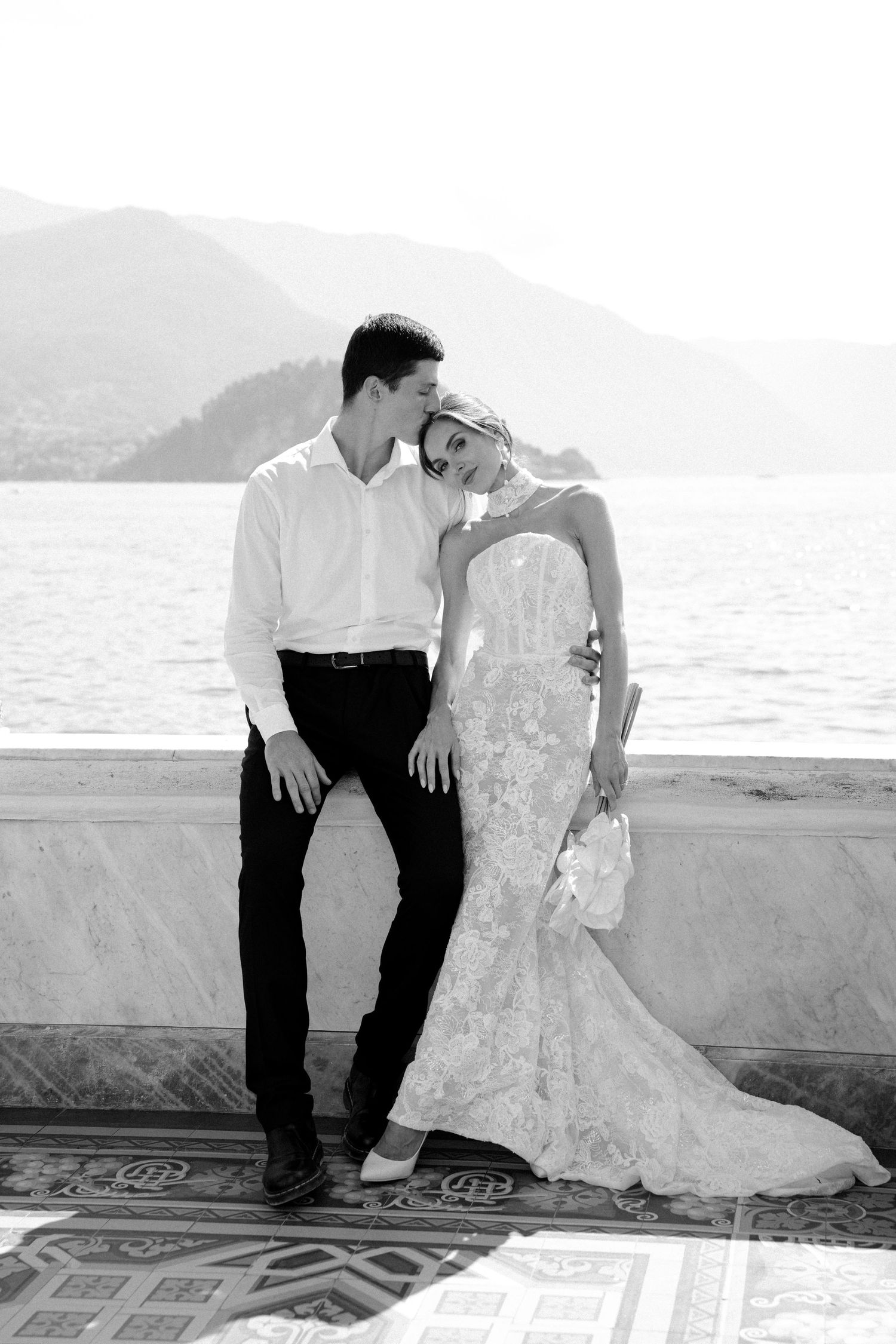 Black and white photo of a bride in lace gown leaning on groom by a lakeside terrace with mountain backdrop.