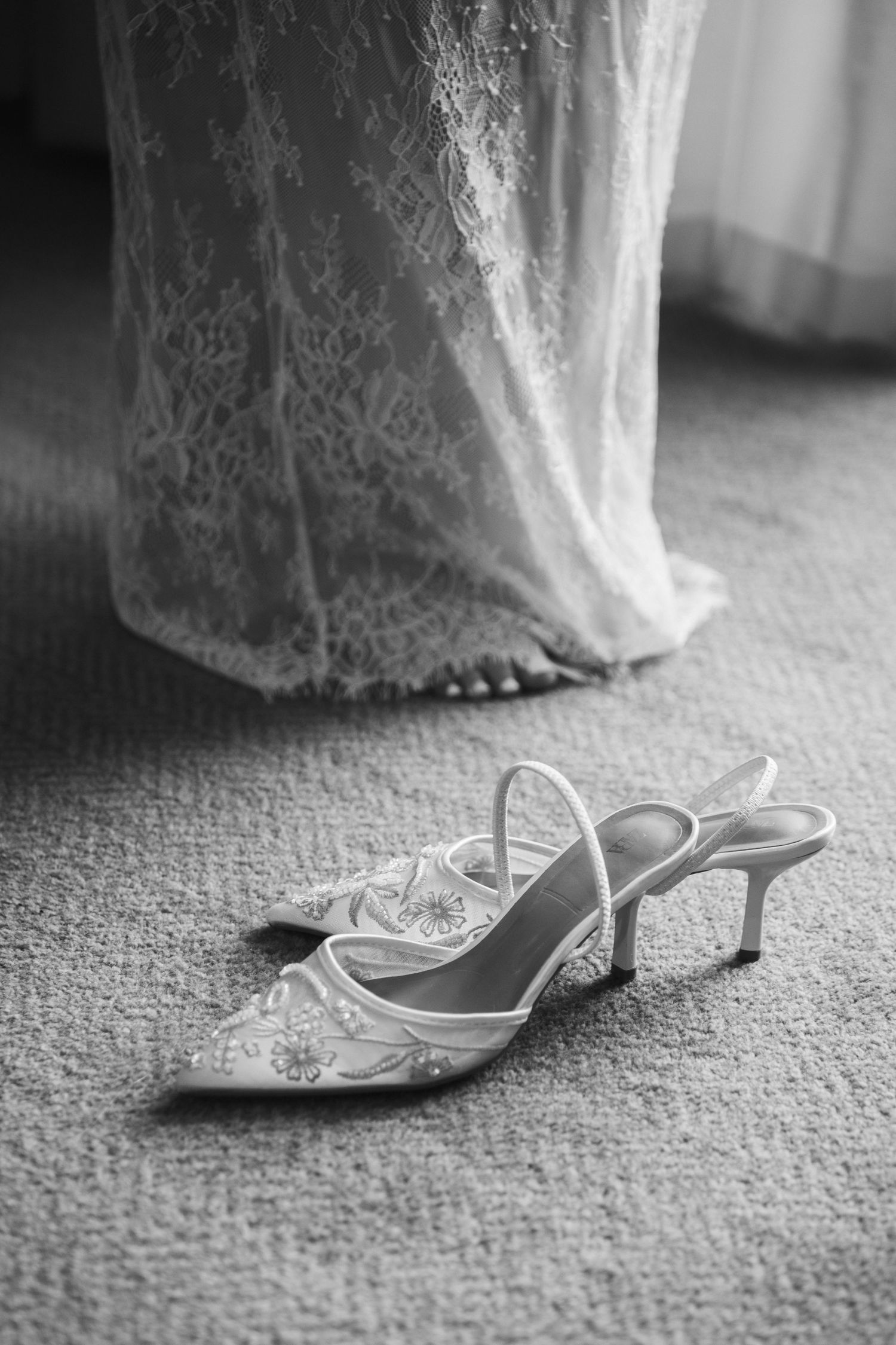 Black and white photo of embellished bridal kitten heel shoes on carpet with lace wedding dress in background.