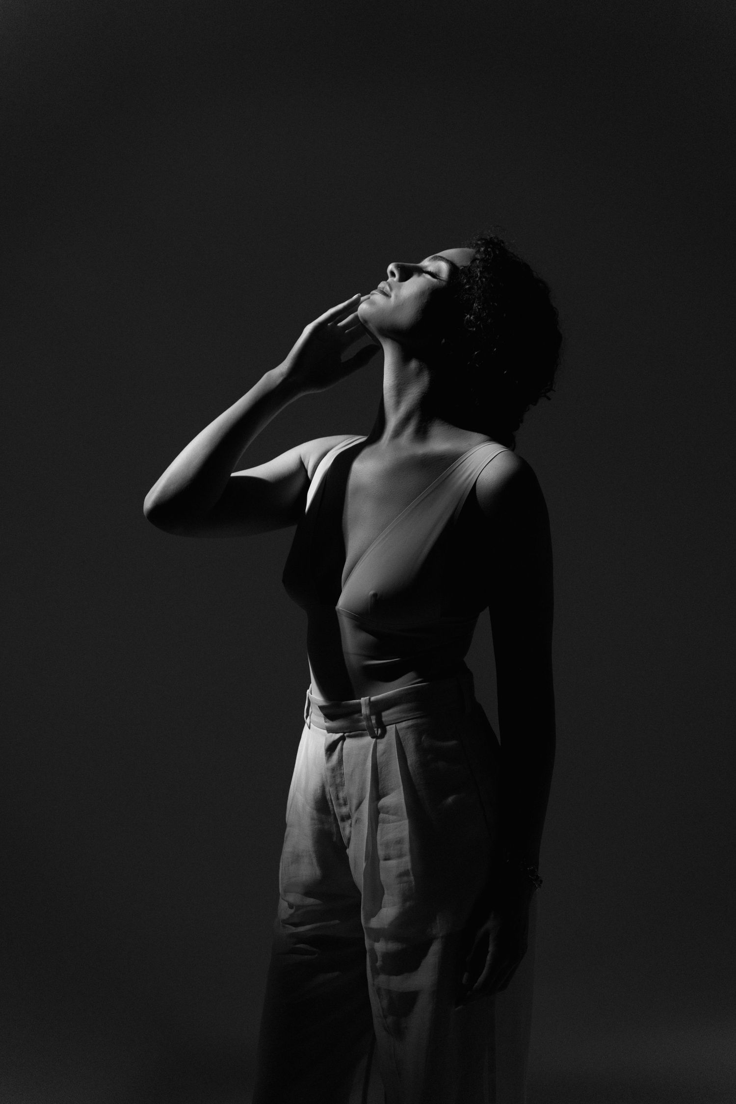Black and white studio portrait of woman in bodysuit and wide-leg pants, head tilted back, dark background.