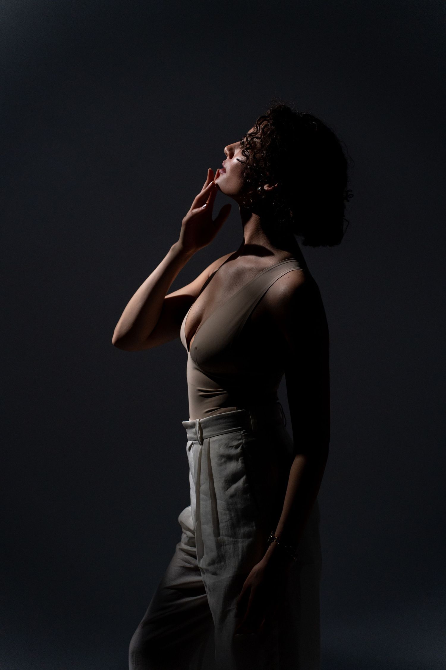 Dramatic low-key portrait of woman in bodysuit and trousers, hand near face, dark studio background.