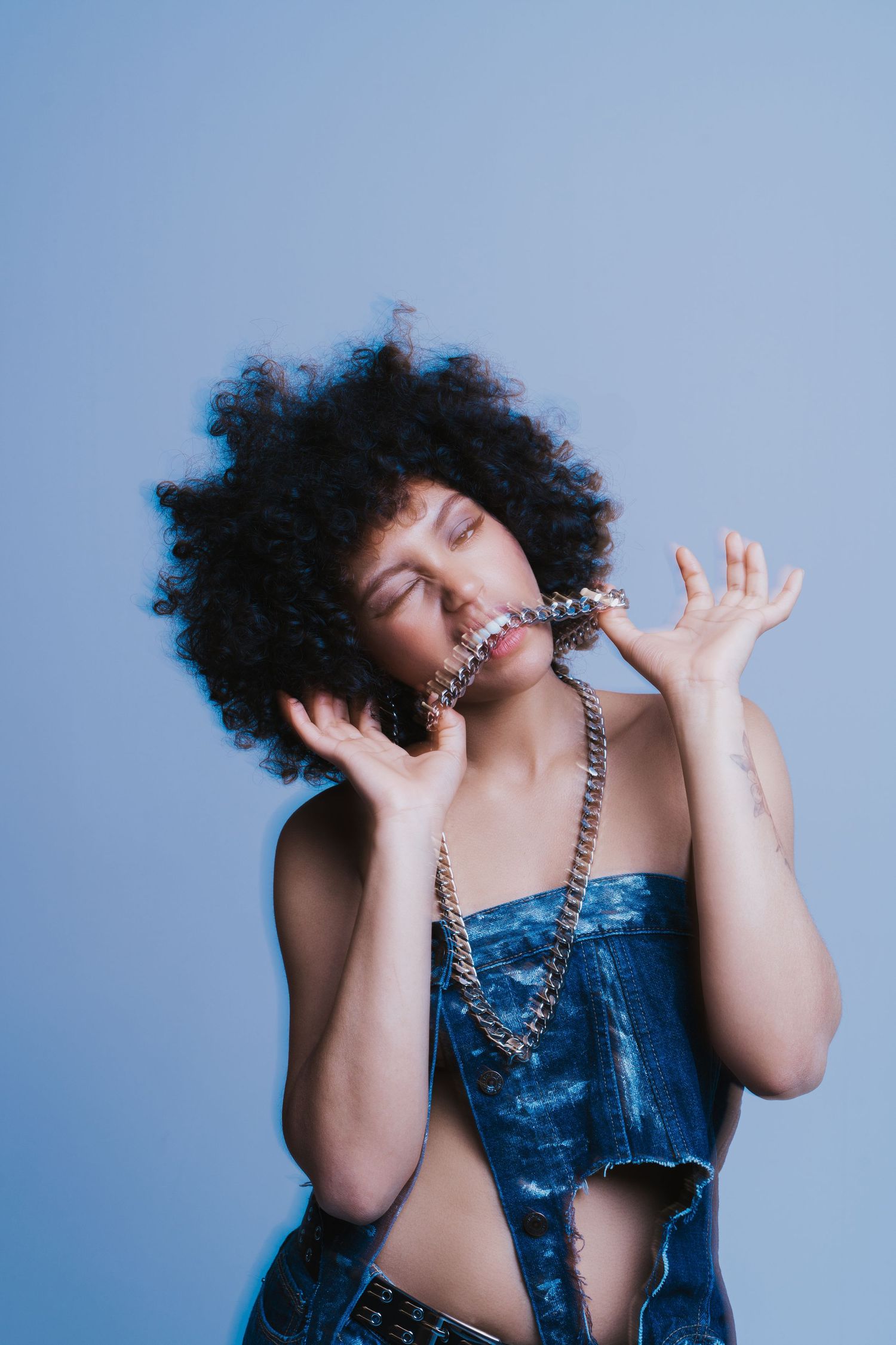 Woman with curly afro hair in denim crop top holding silver chain necklace on blue background.