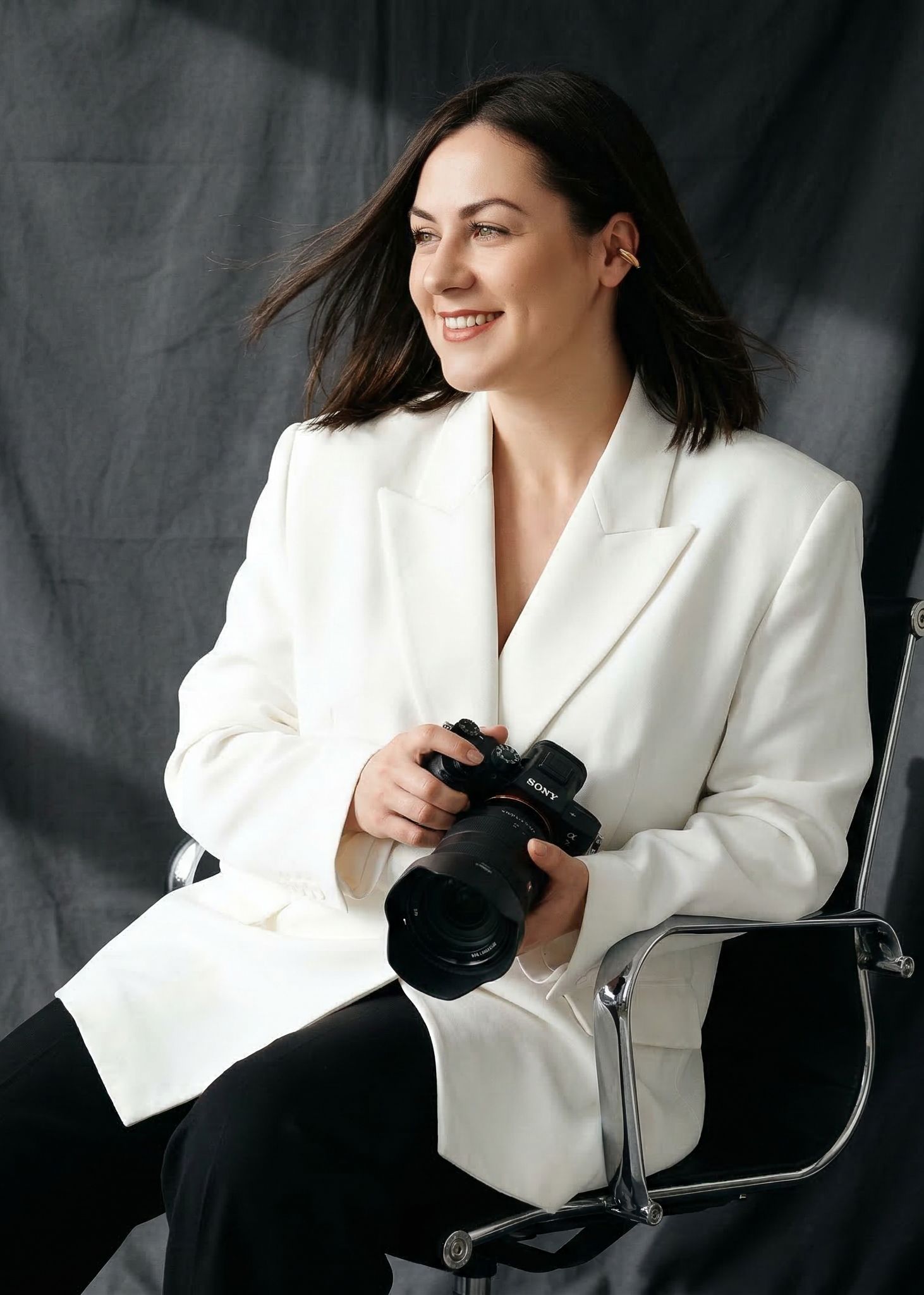 Smiling dark-haired female photographer in a white blazer holding a Sony camera while seated in a chair.
