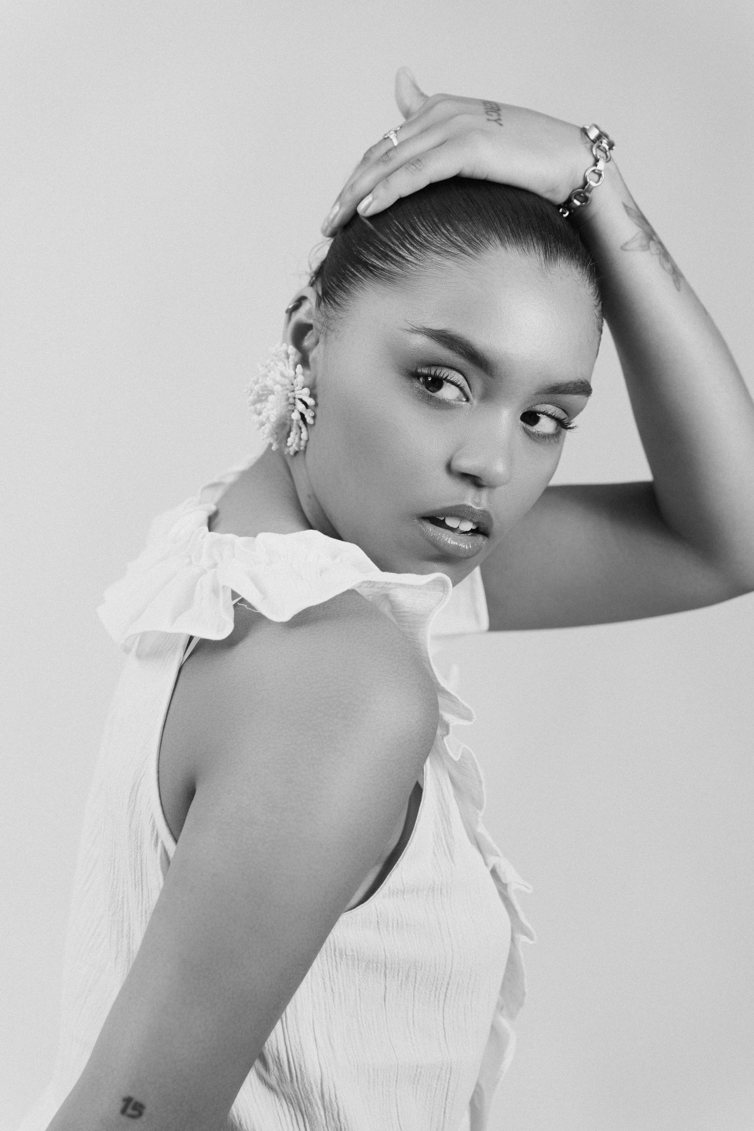 Black and white studio portrait of a woman in a white ruffled top, hand on head, wearing floral earrings.