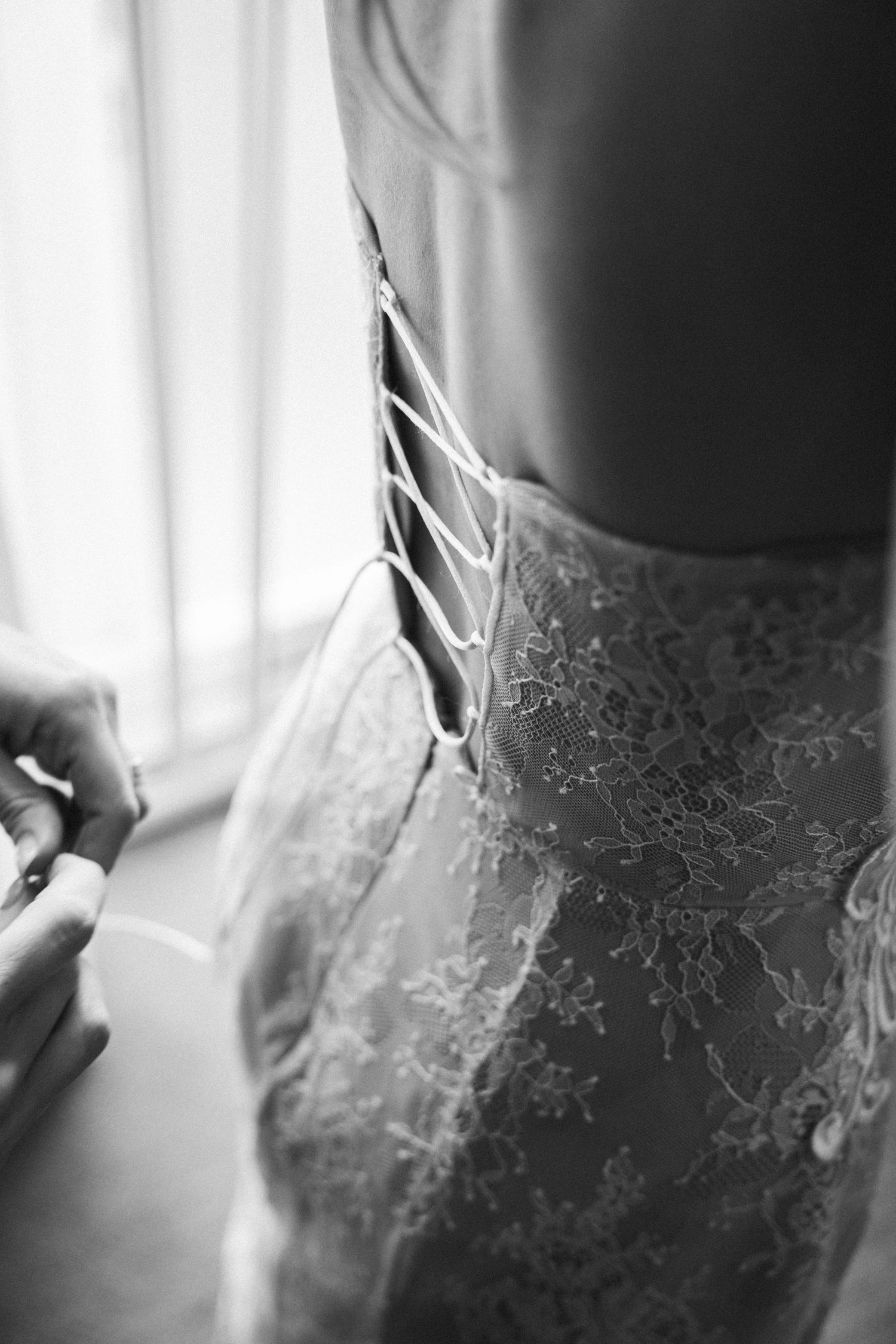Close-up black and white detail of a bride's lace corset wedding dress being laced up before ceremony.