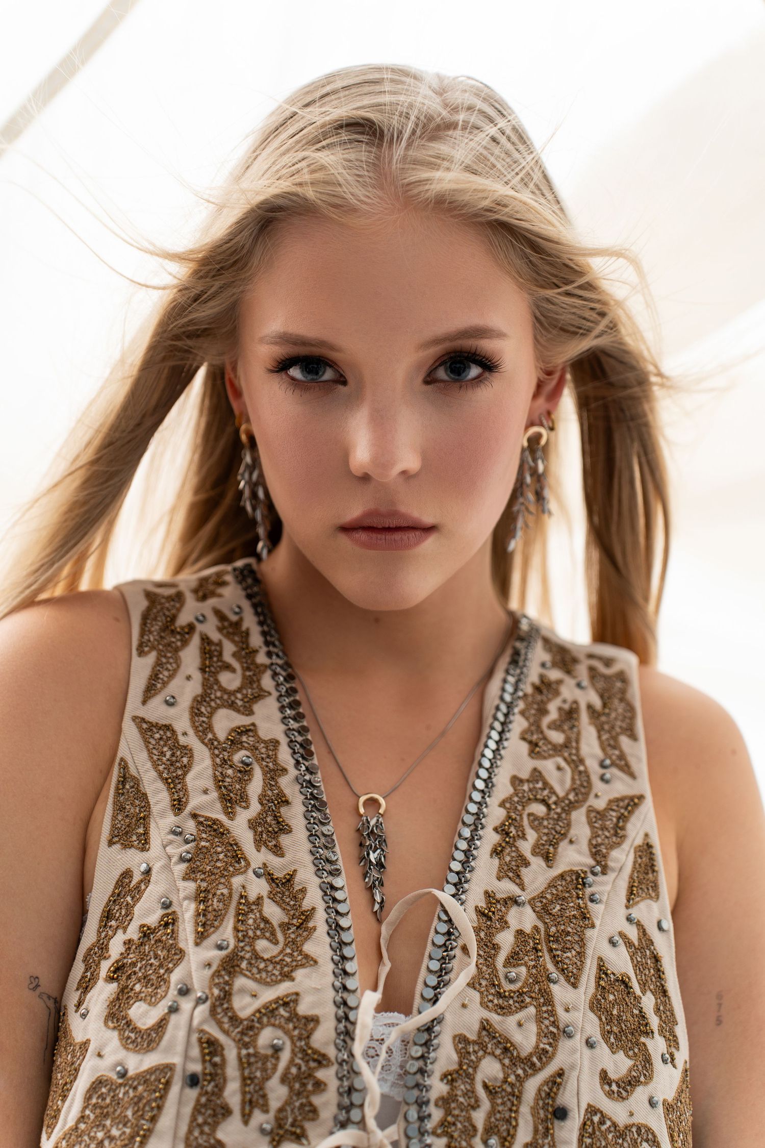 Close-up portrait of a blonde woman wearing layered necklaces and embroidered top with earrings.