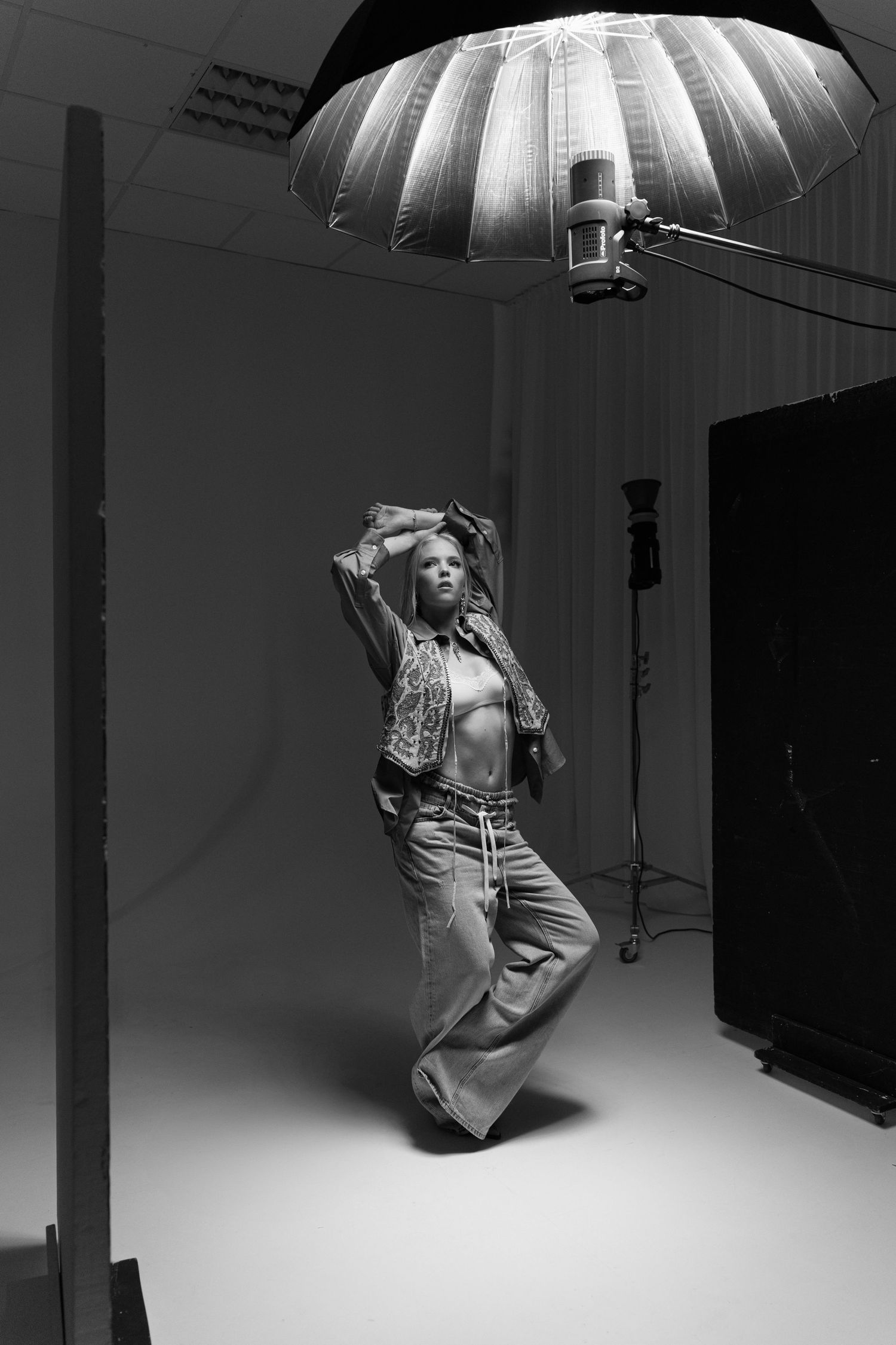 Black and white behind-the-scenes studio photo of a model posing under a large umbrella light.