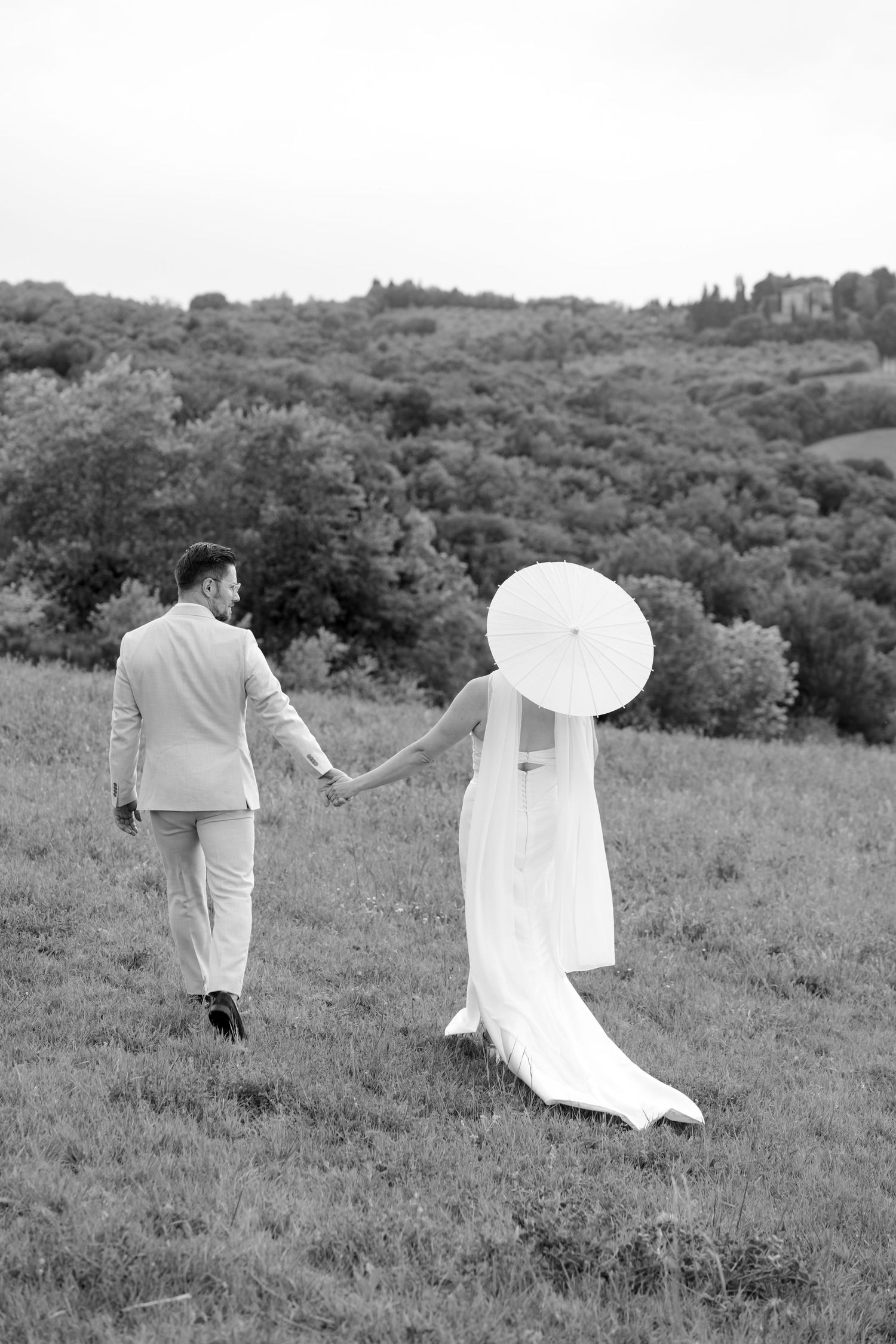 Black and white image of a bride with parasol and groom walking hand in hand across a scenic green hillside.