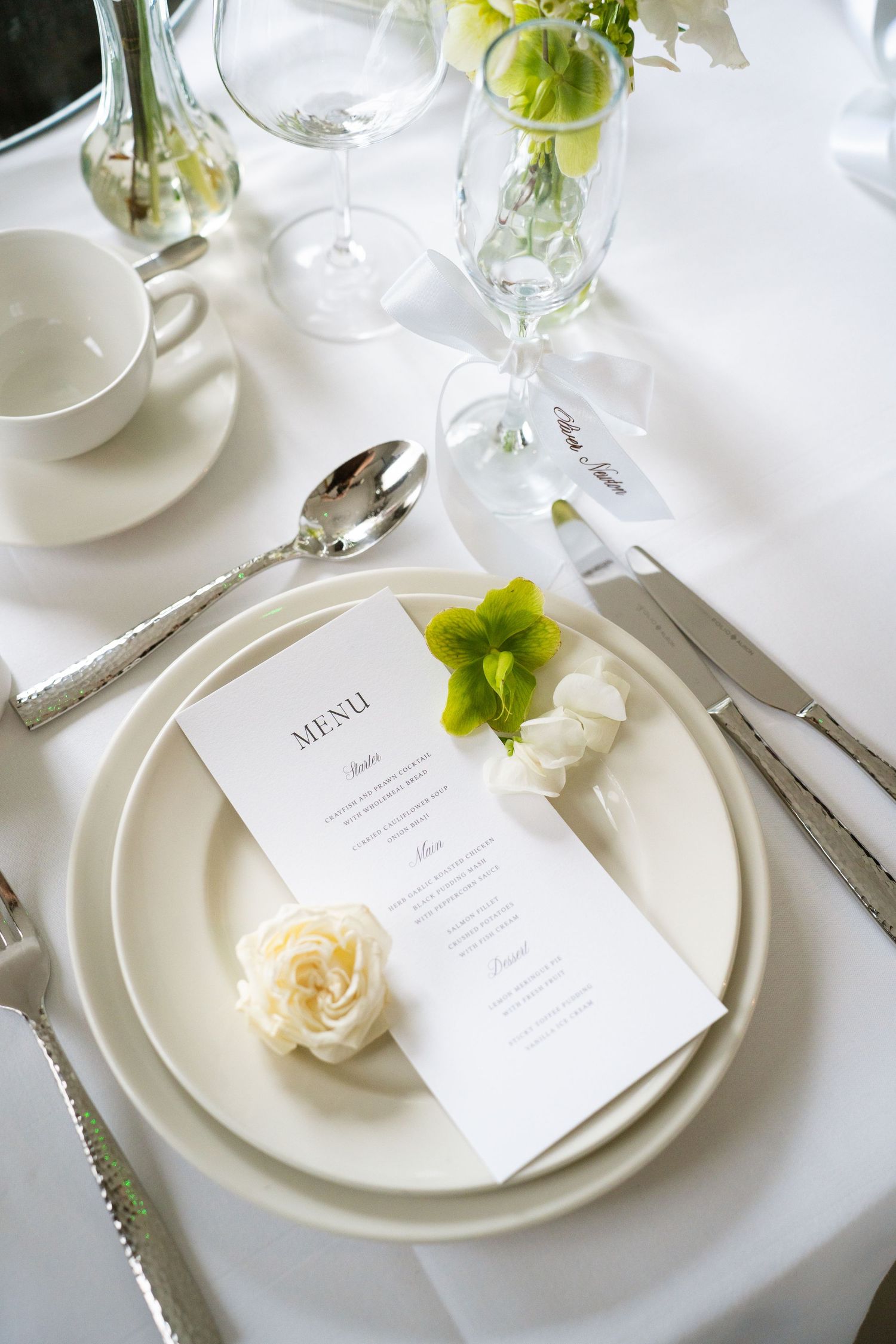 Elegant wedding table setting with white plates, silver cutlery, menu card, and fresh flowers on a white tablecloth.