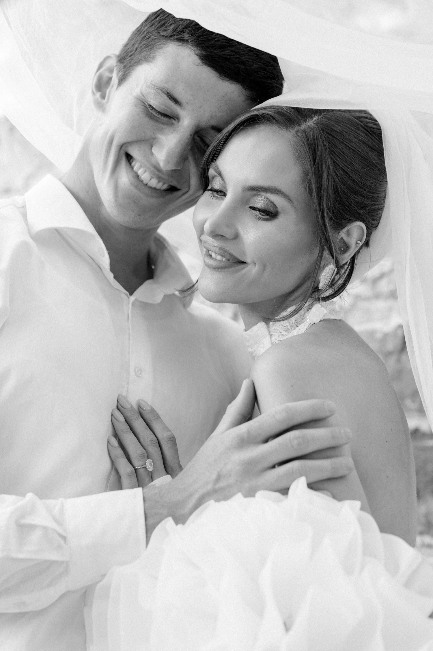 Black and white wedding photo of smiling bride and groom embracing under a veil, bride wearing engagement ring.