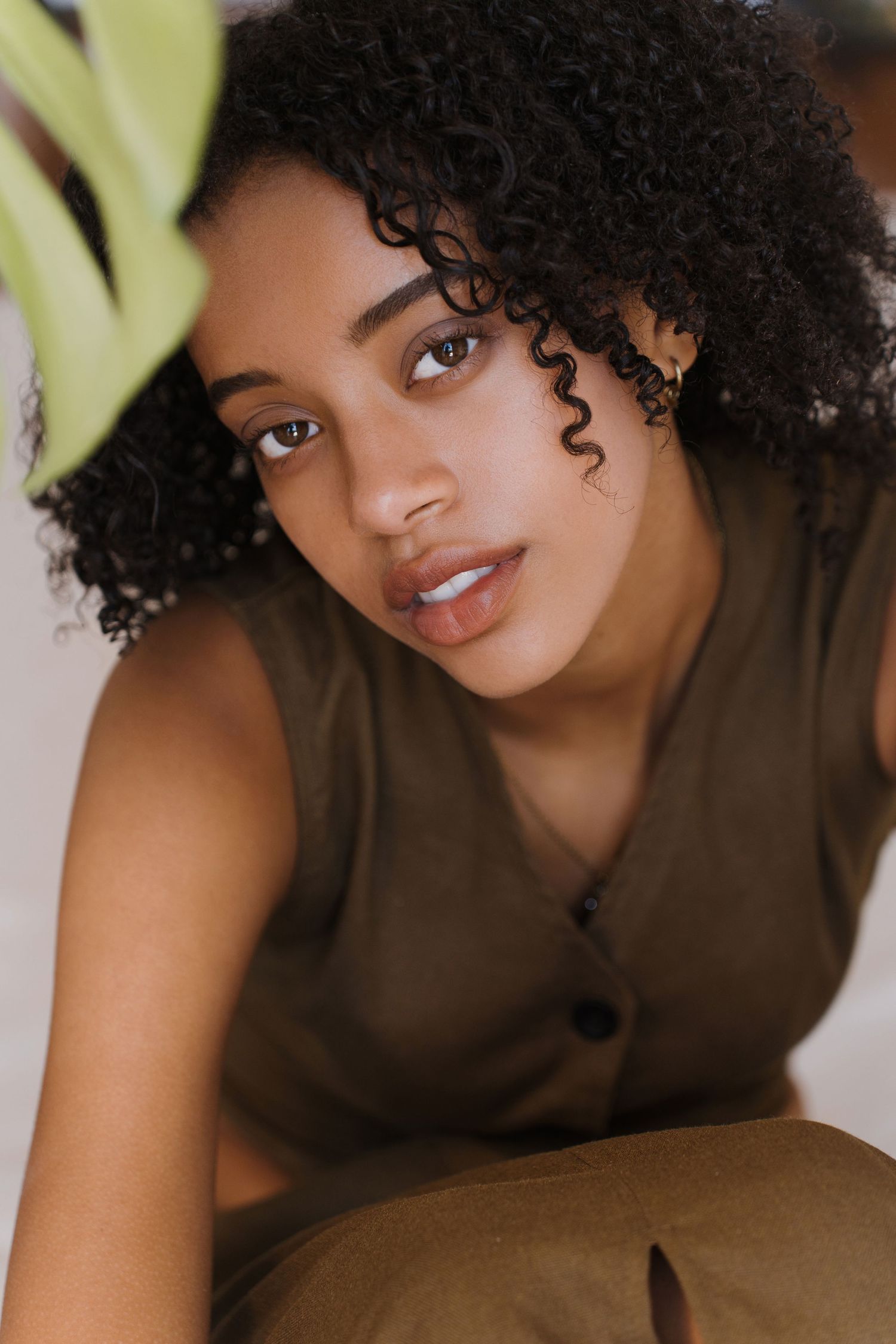 Young Black woman with natural curly hair wearing olive sleeveless top, gazing at camera in soft natural light.