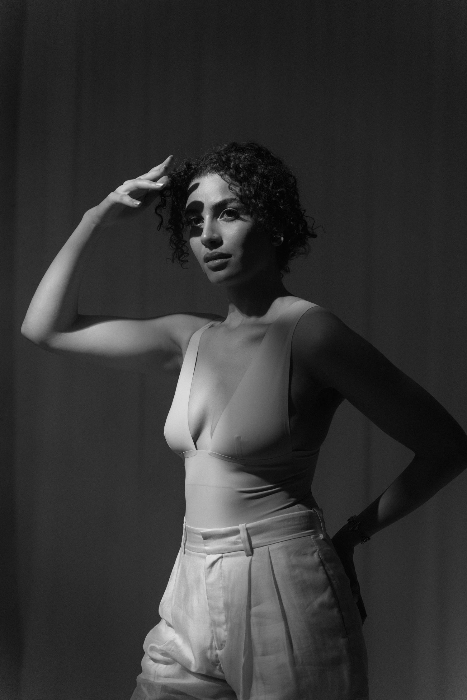 Black and white portrait of curly-haired woman in bodysuit and trousers, hand raised to forehead.