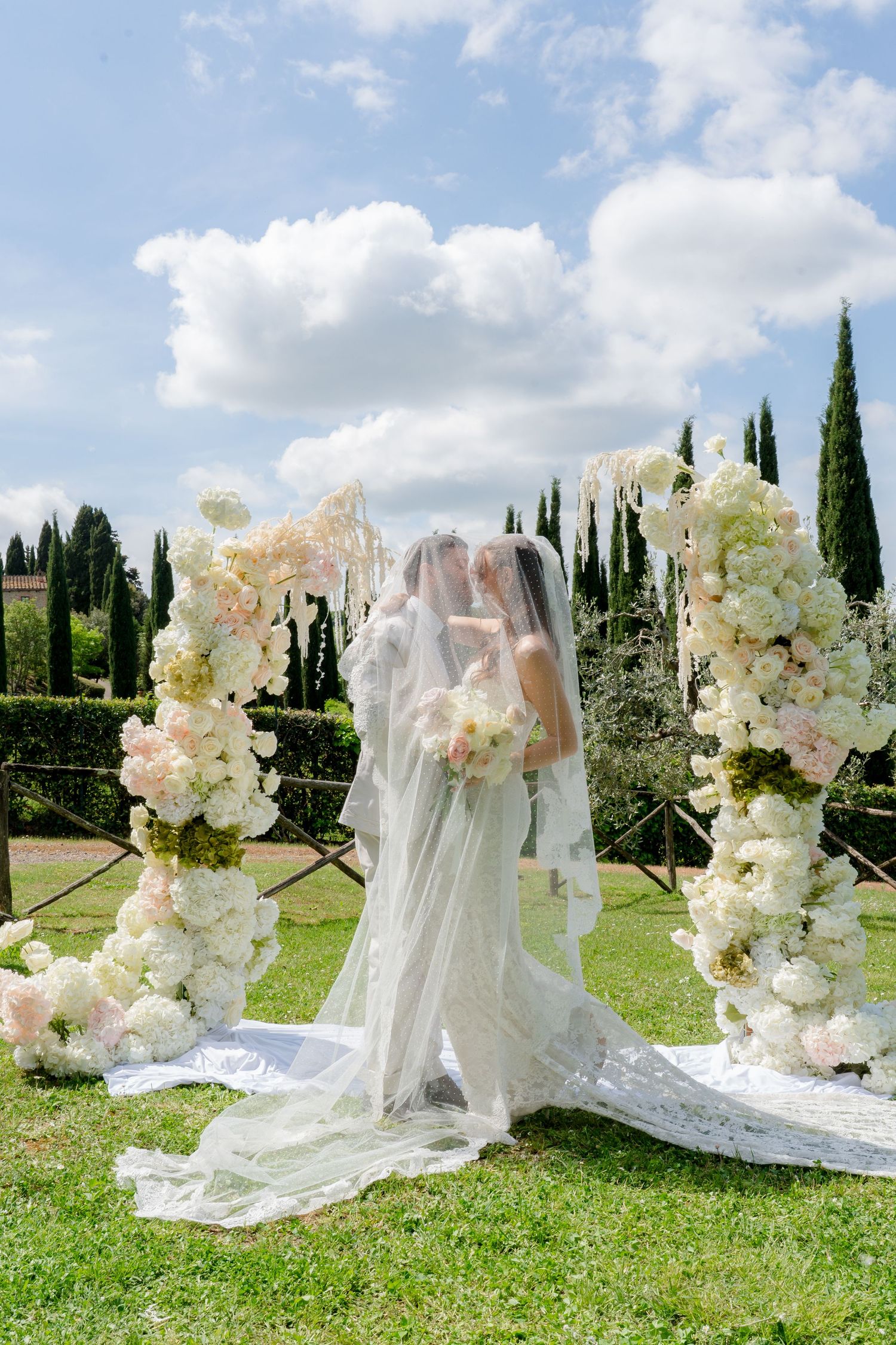 Two brides share a kiss beneath a floral arch of white and blush blooms at an outdoor ceremony.