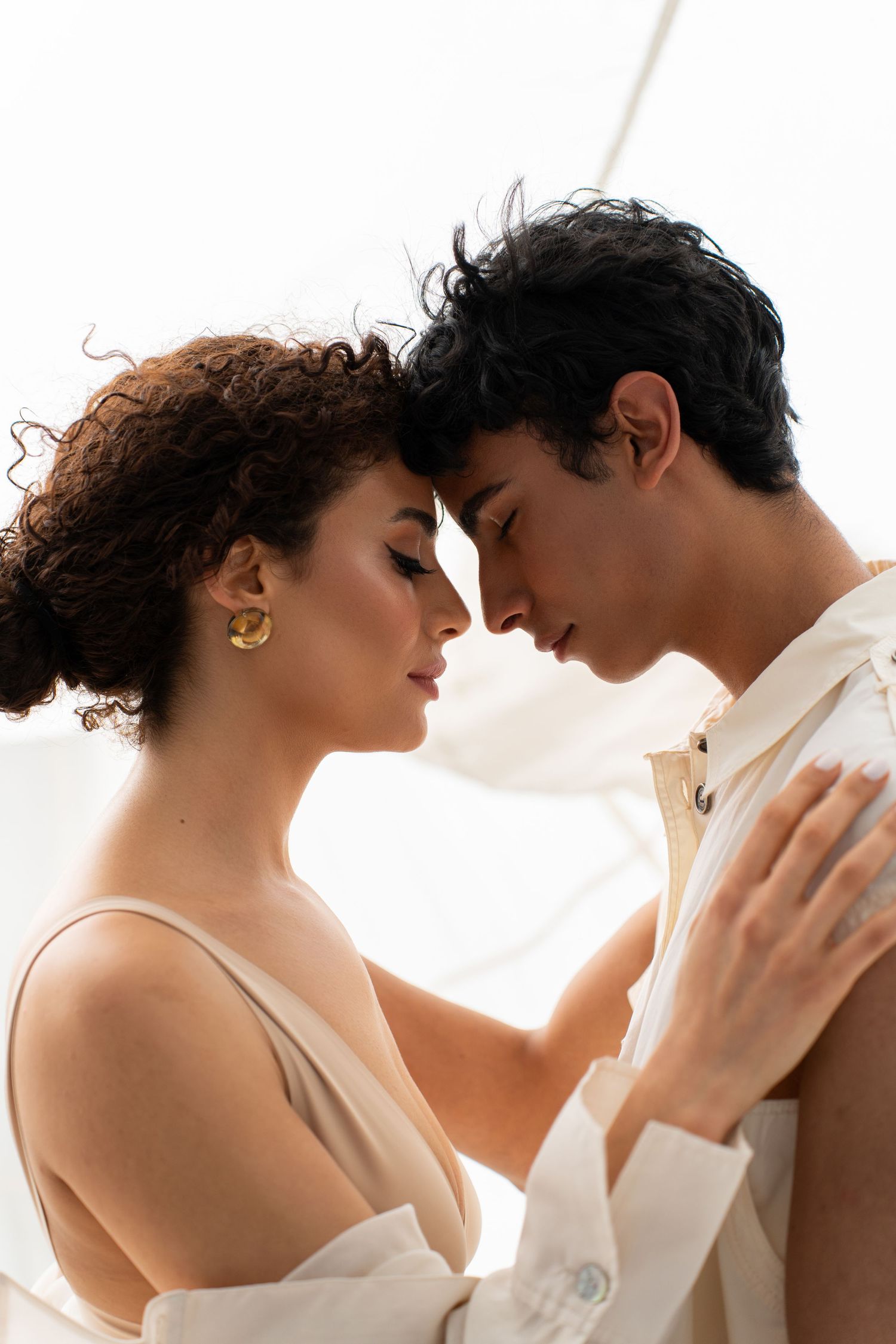 Romantic couple with curly hair embracing closely, wearing white outfits against a light background.