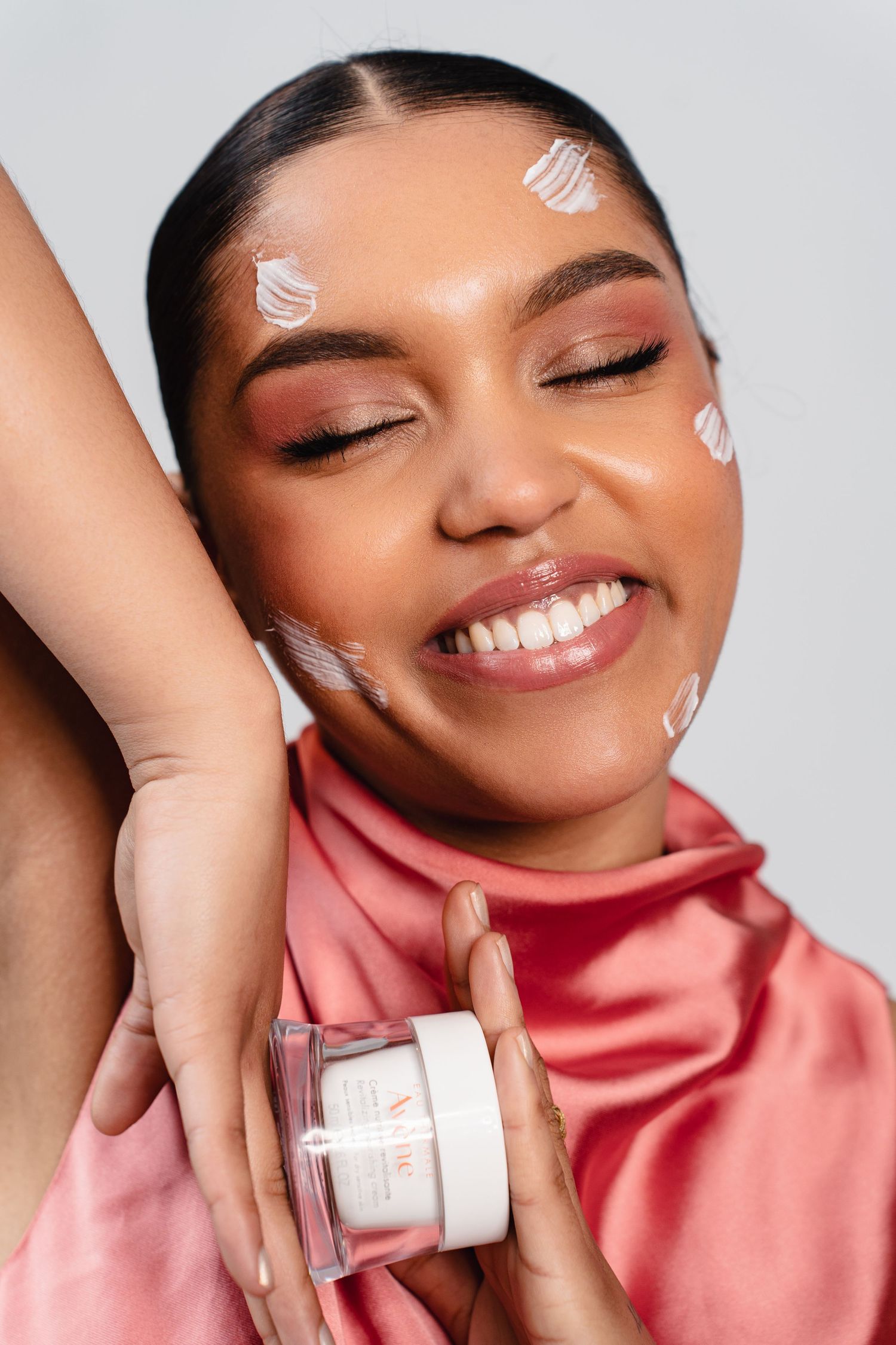 Smiling woman with closed eyes in pink satin top holds skincare cream jar with white product dots on her face.